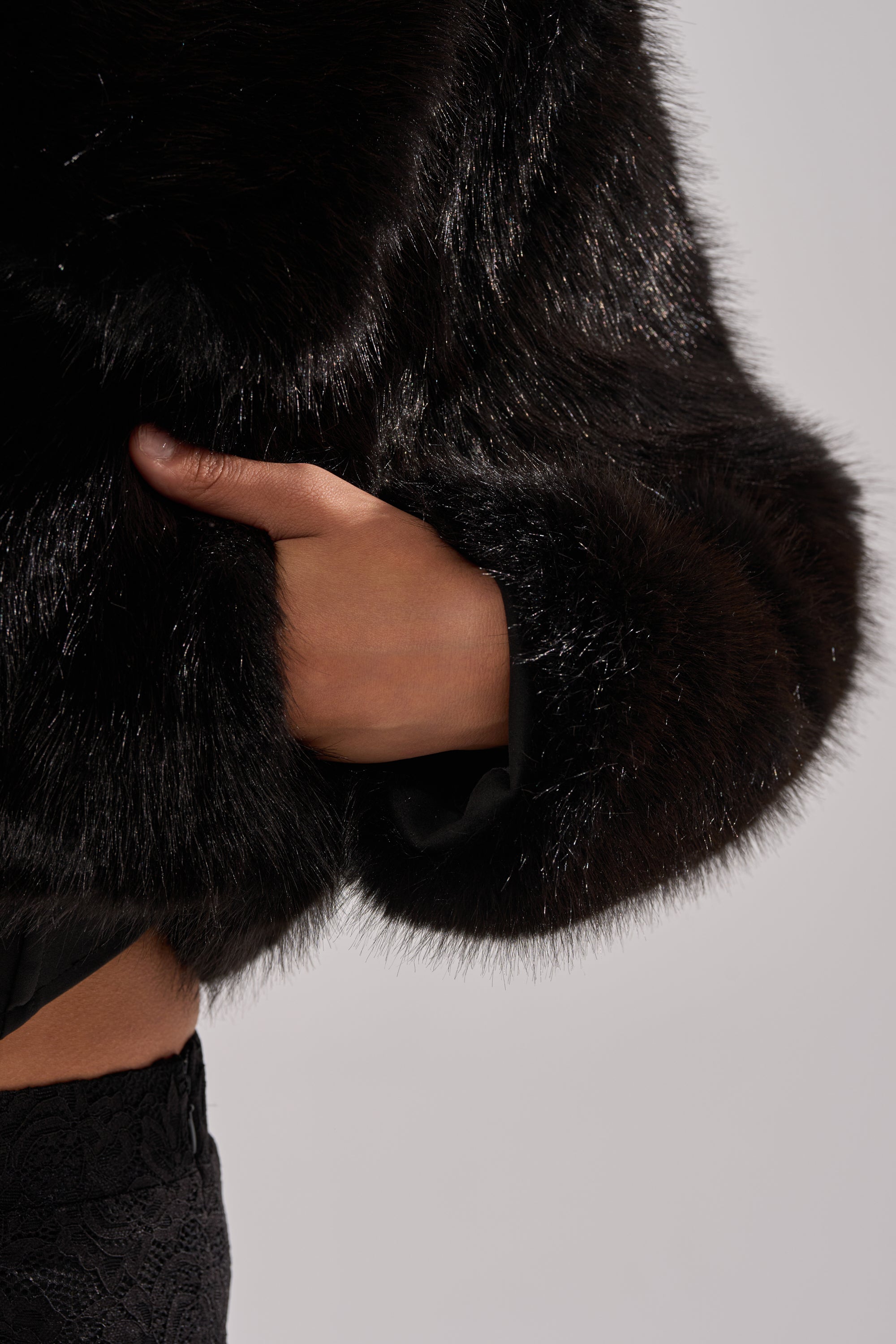 A hand rests in the pocket of the CADENCE FAUX FUR IN BLACK coat, paired with textured black pants, set against a simple light background.