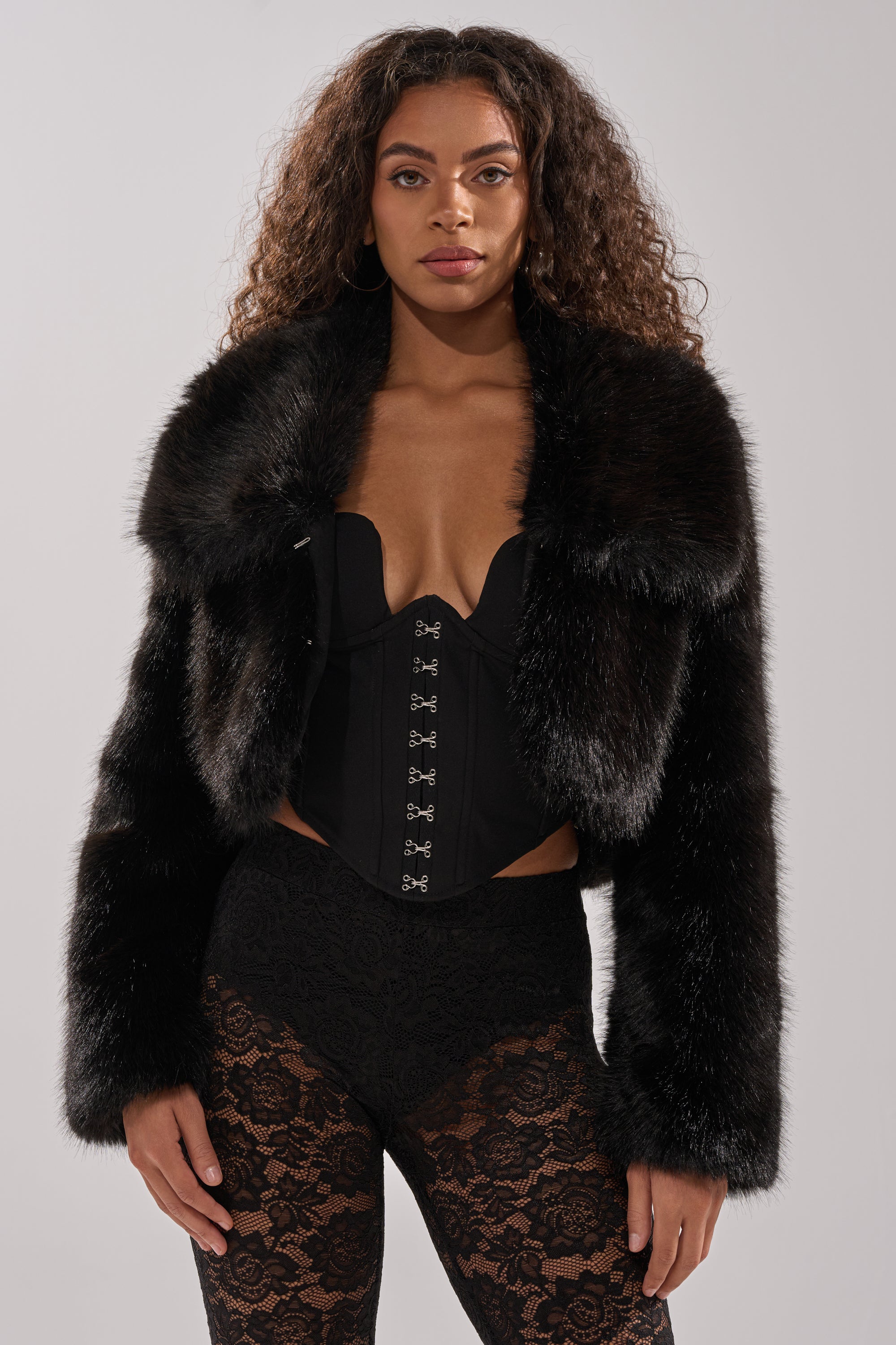A woman with long curly hair models the CADENCE FAUX FUR IN BLACK over a black corset top and lace pants, standing against a plain light gray backdrop.