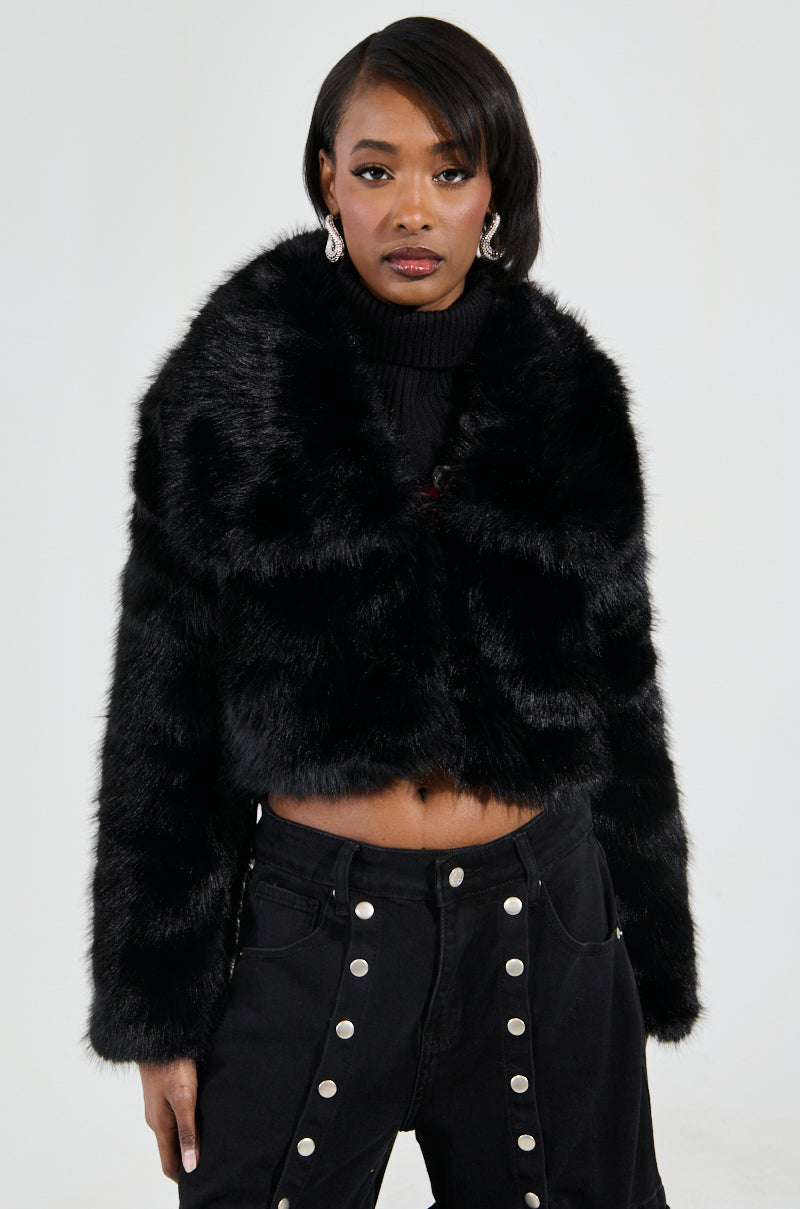 A person stands confidently against a plain background, wearing the CADENCE FAUX FUR IN BLACK coat paired with high-waisted black pants featuring decorative buttons.