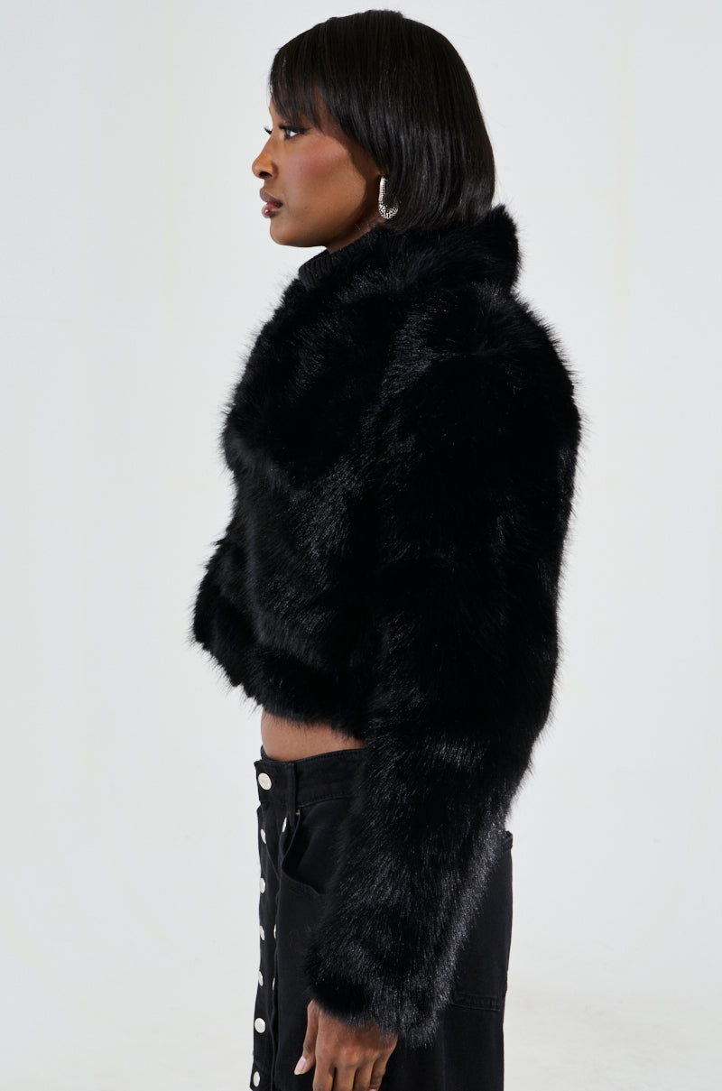 A person with shoulder-length dark hair stands in profile against a plain background, showcasing their style in fashionable outerwear. They confidently wear the CADENCE FAUX FUR IN BLACK coat paired with matching black pants featuring white buttons, as they gaze to the right.