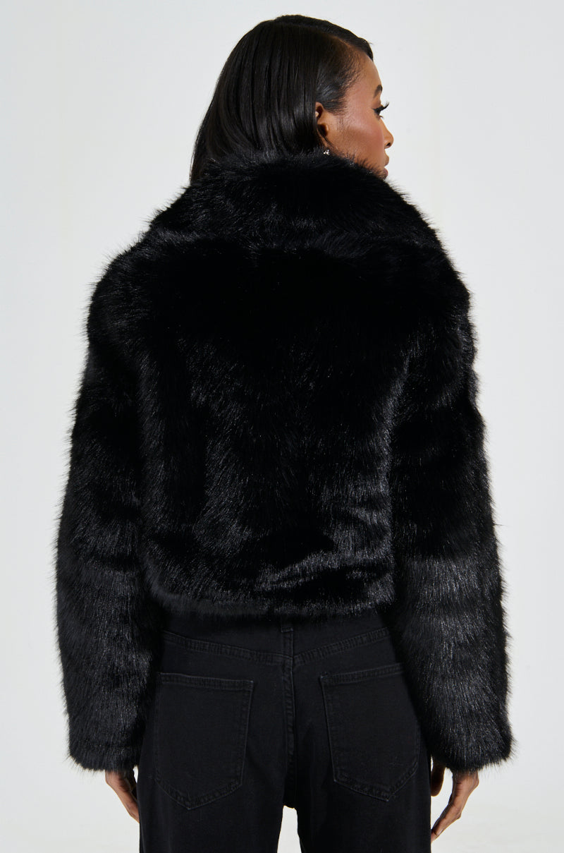 A person with long dark hair is seen from behind, wearing the fashionable CADENCE FAUX FUR IN BLACK coat and matching pants. The plain, light-colored background accentuates the luxurious texture of the outerwear.