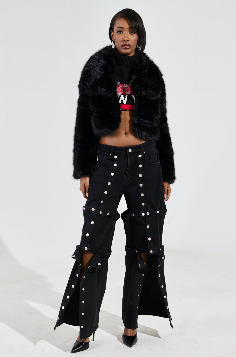 A woman exudes confidence as she poses on a plain background, draped in the CADENCE FAUX FUR IN BLACK from AZALEA WANG. She wears this stunning faux fur coat over a cropped top and black pants adorned with white buttons, finishing the look with stylish high heels.