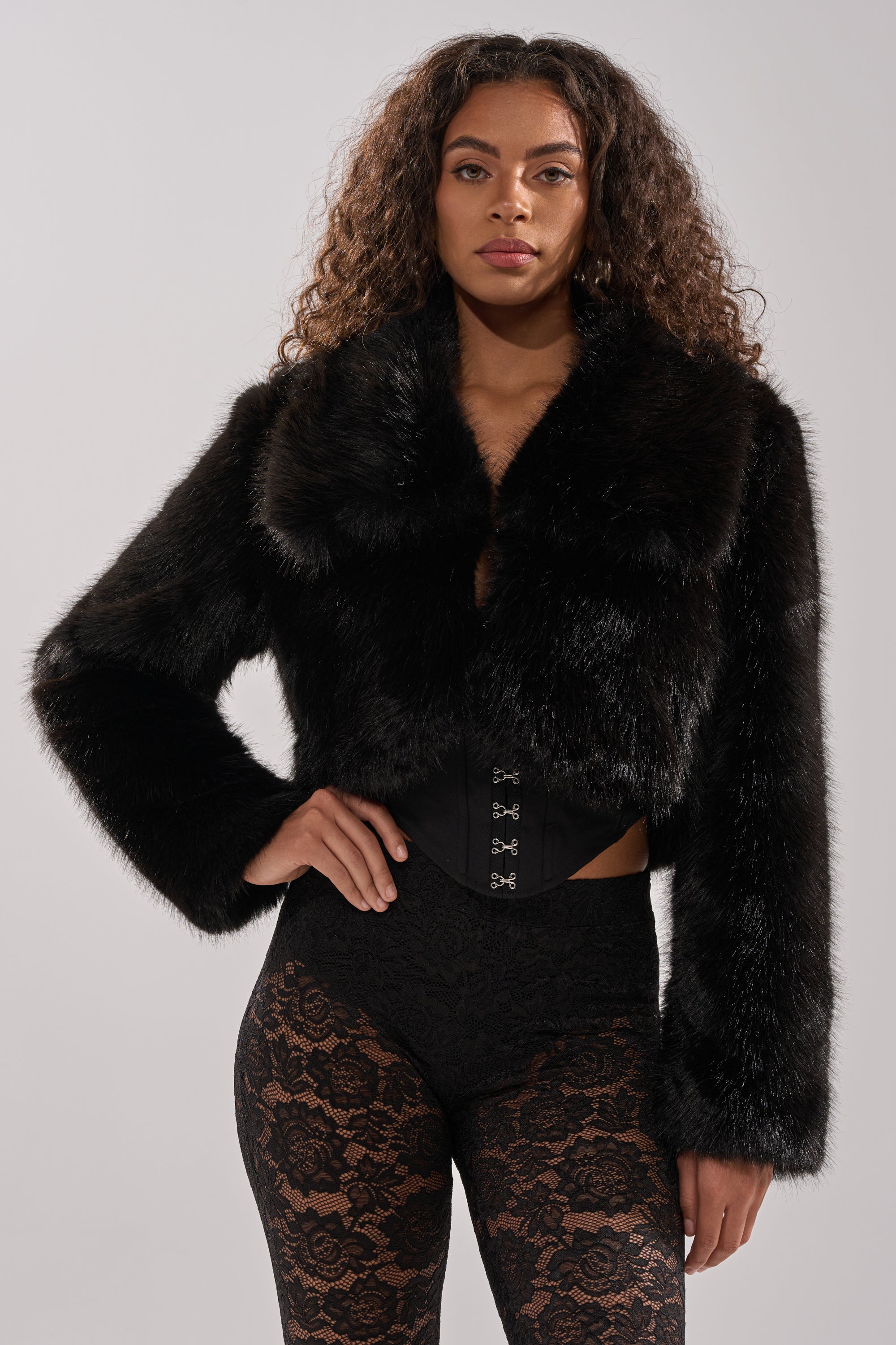 A woman with long curly hair models the CADENCE FAUX FUR IN BLACK over a black corset and sheer black lace pants, standing against a plain light gray background.