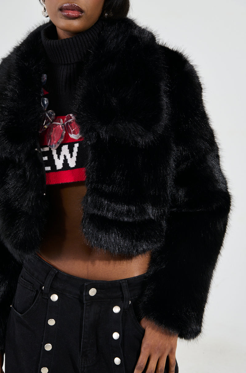 A person wearing the CADENCE FAUX FUR IN BLACK over a red and black cropped sweater with text and a beaded necklace. Their pants are black with white buttons, completing an overall stylish and modern look.