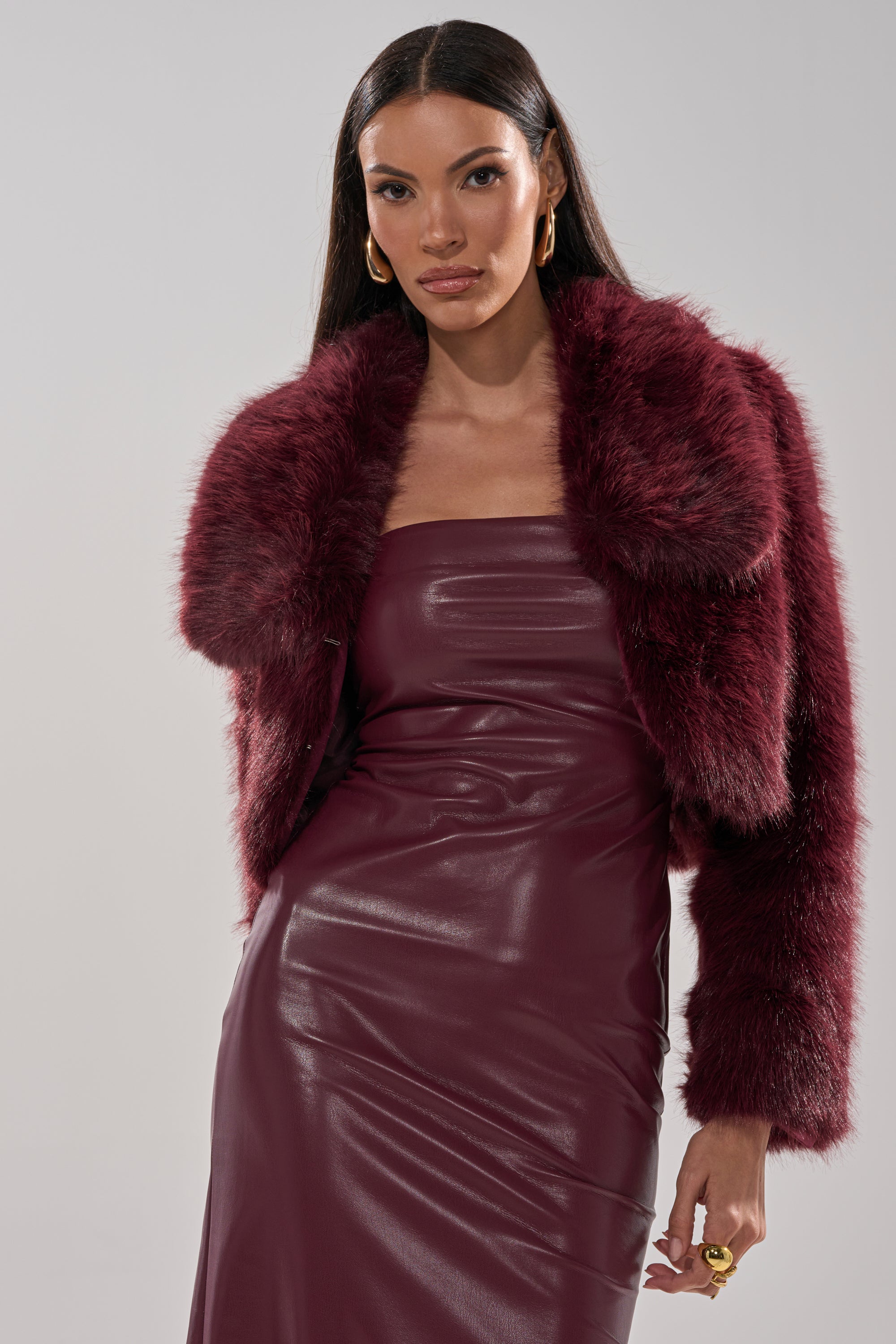 A woman in the CADENCE FAUX FUR IN BURGUNDY jacket wears a strapless fitted leather dress, gold hoop earrings, and has long dark hair. She poses confidently against a plain light background.