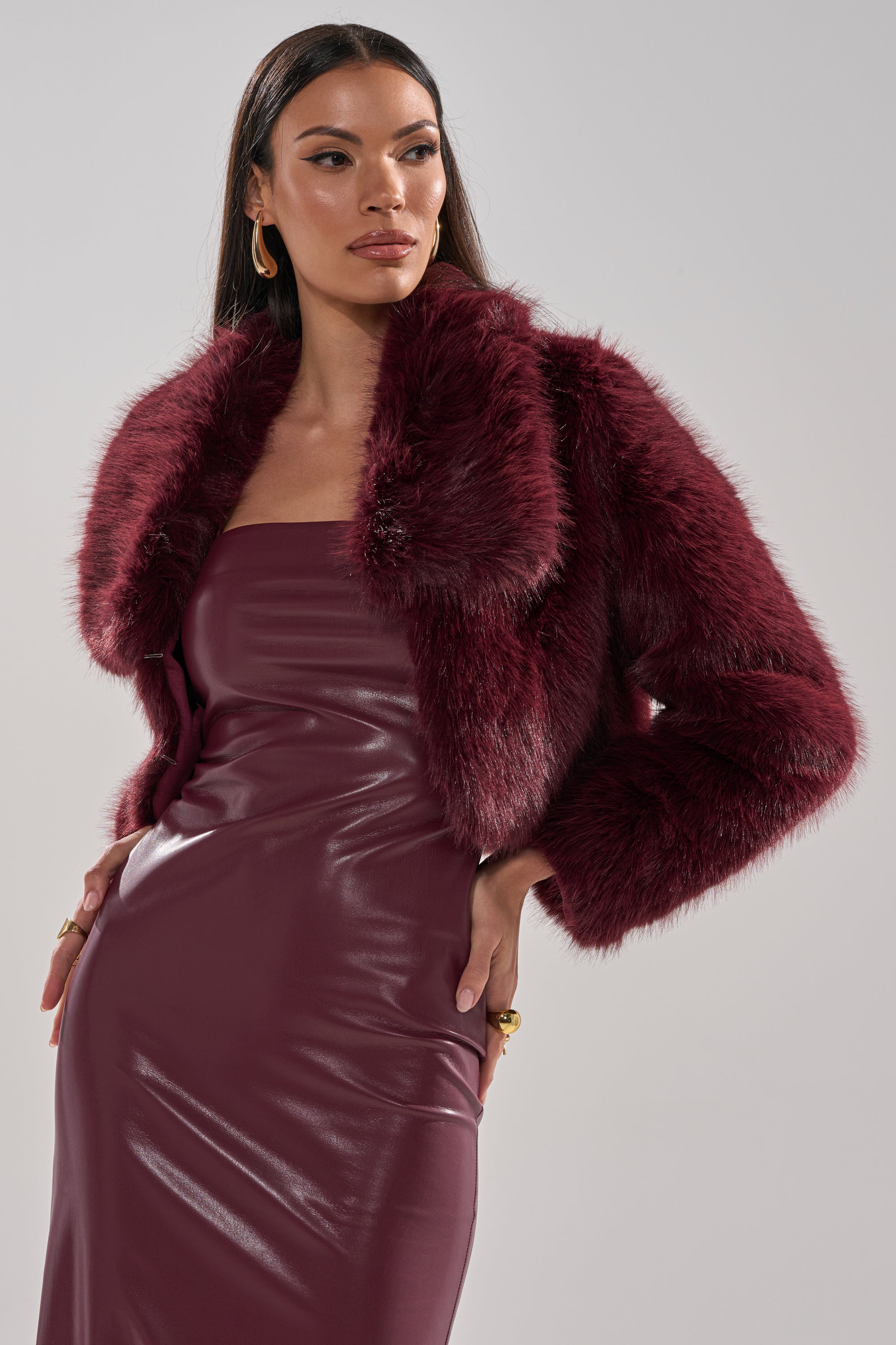 A woman models the CADENCE FAUX FUR IN BURGUNDY, styled over a sleeveless, fitted burgundy leather dress. She accessorizes with gold hoop earrings and rings, her long hair straight, standing against a simple light background.