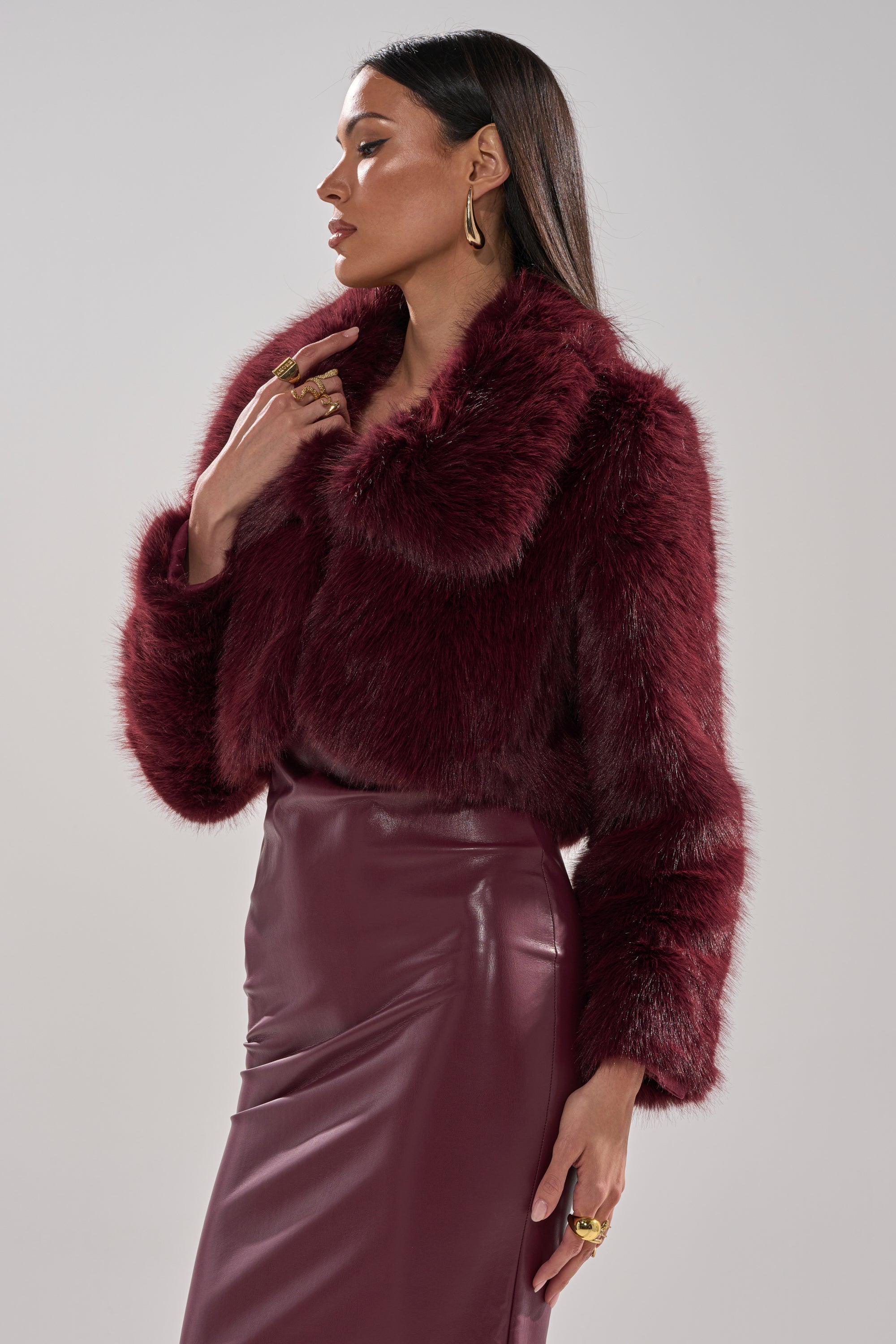 A woman stands in profile against a plain background, wearing the CADENCE FAUX FUR IN BURGUNDY jacket and a matching skirt, with her left hand raised and her head turned slightly to the side.