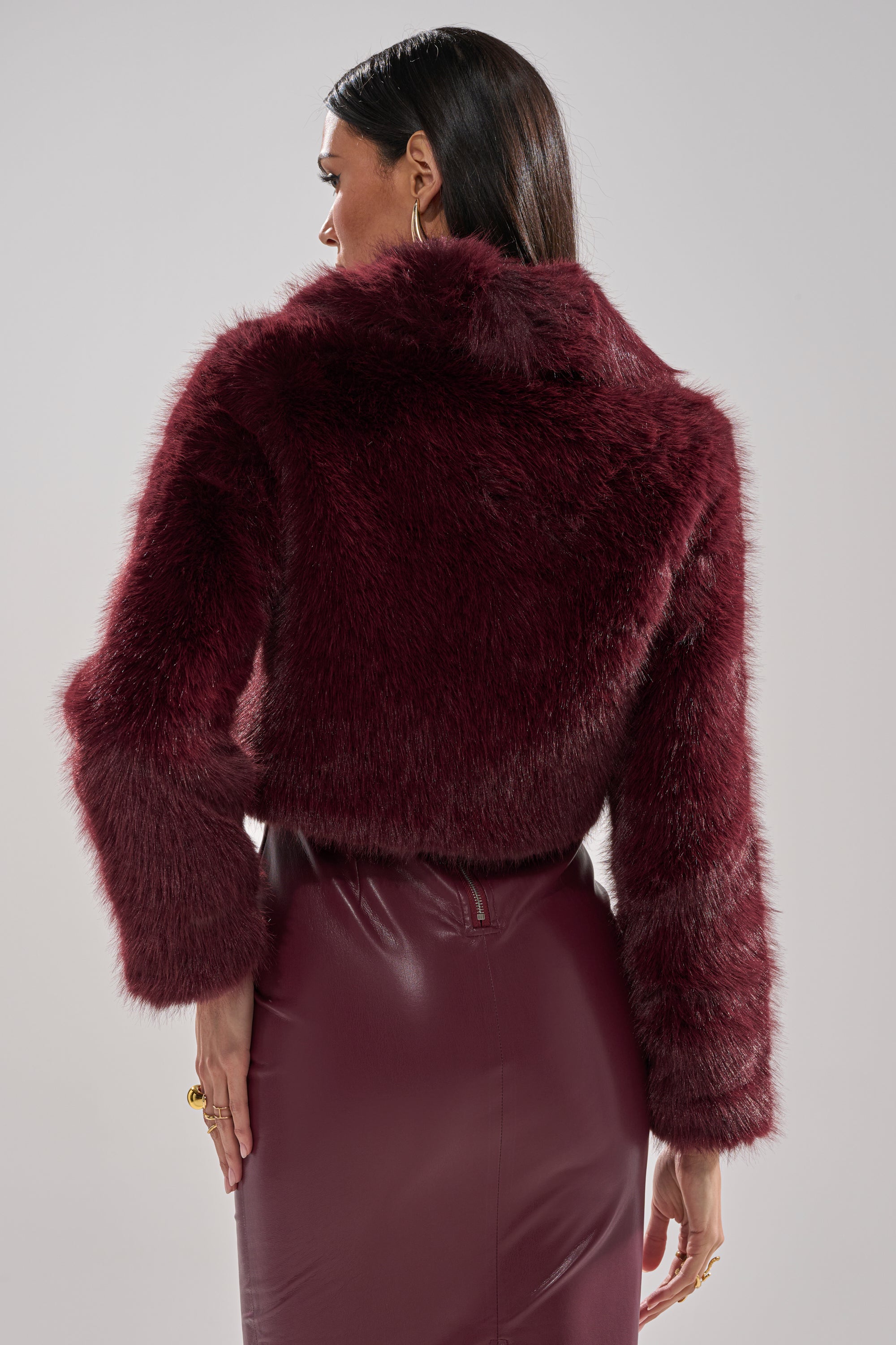 A person with long dark hair, seen from behind against a neutral background, showcases the elegance of the CADENCE FAUX FUR IN BURGUNDY—styled with a matching high-waisted leather skirt for a refined look.