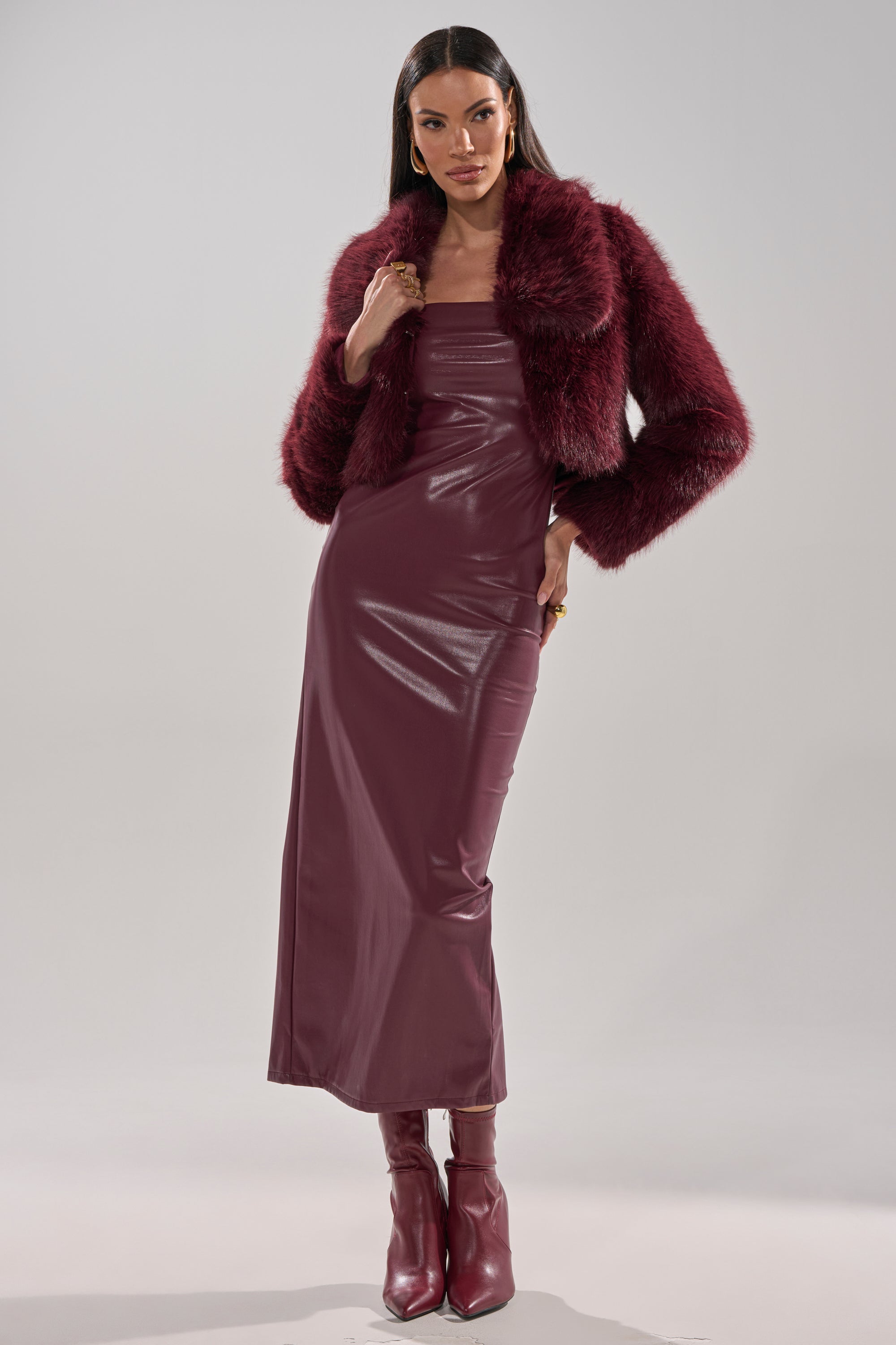 A woman models the CADENCE FAUX FUR IN BURGUNDY jacket over a matching strapless, form-fitting, ankle-length dress with burgundy heeled boots, set against a plain light gray background.
