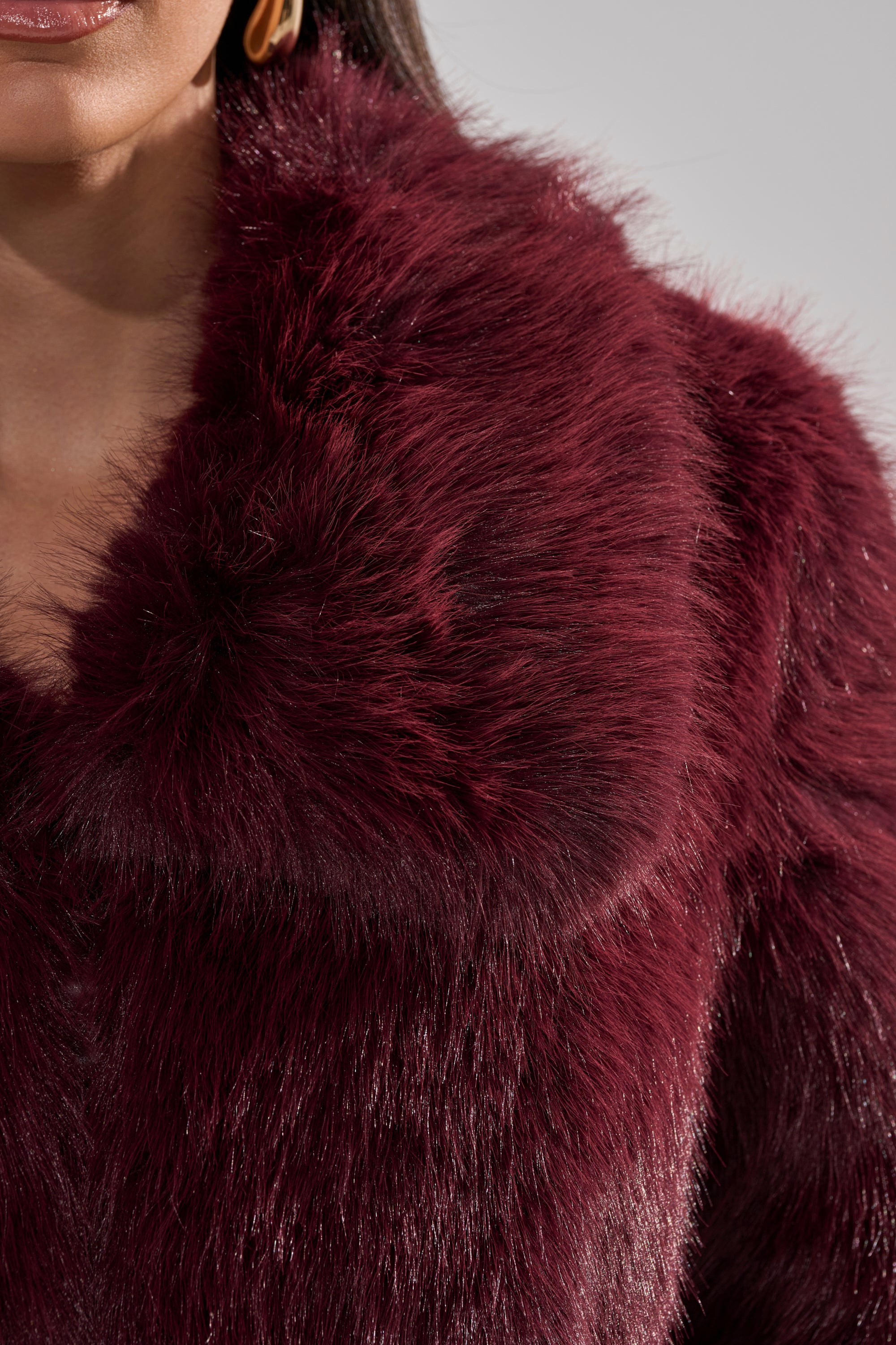 Close-up of a person wearing the CADENCE FAUX FUR IN BURGUNDY, showcasing its rich texture and sheen. The image captures the lower face and a gold hoop earring, adding an elegant touch.