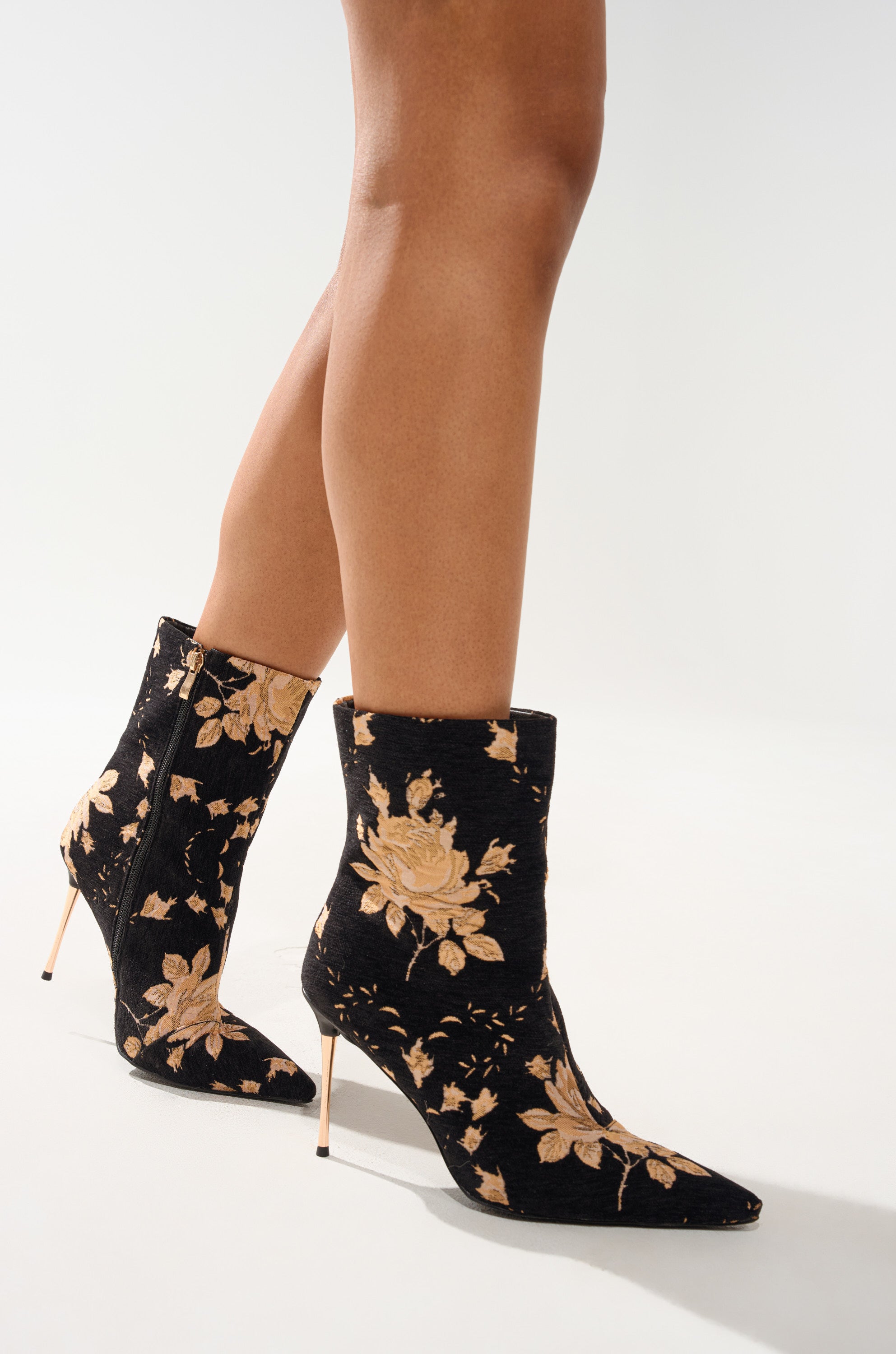 A person stands on a light background wearing the AZALEA WANG CALEAN BLACK MULTI BROCADE STILETTO BOOTIE, featuring black floral brocade with gold patterns, pointed toes, and thin high heels. 0