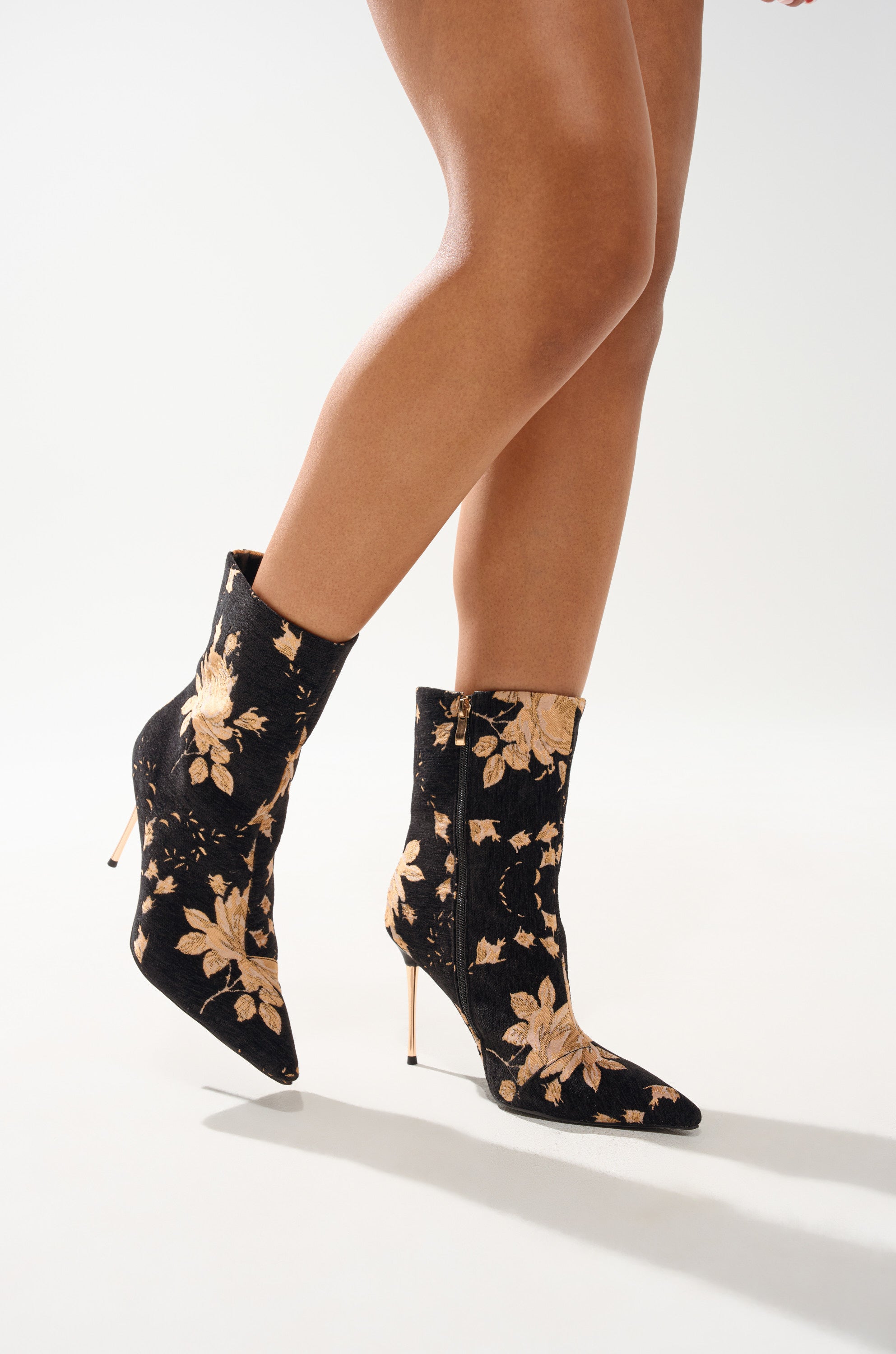 A person wears the AZALEA WANG CALEAN BLACK MULTI BROCADE STILETTO BOOTIE, featuring black floral brocade with gold patterns and pointed toes, shown from mid-thigh down against a plain light background. 2