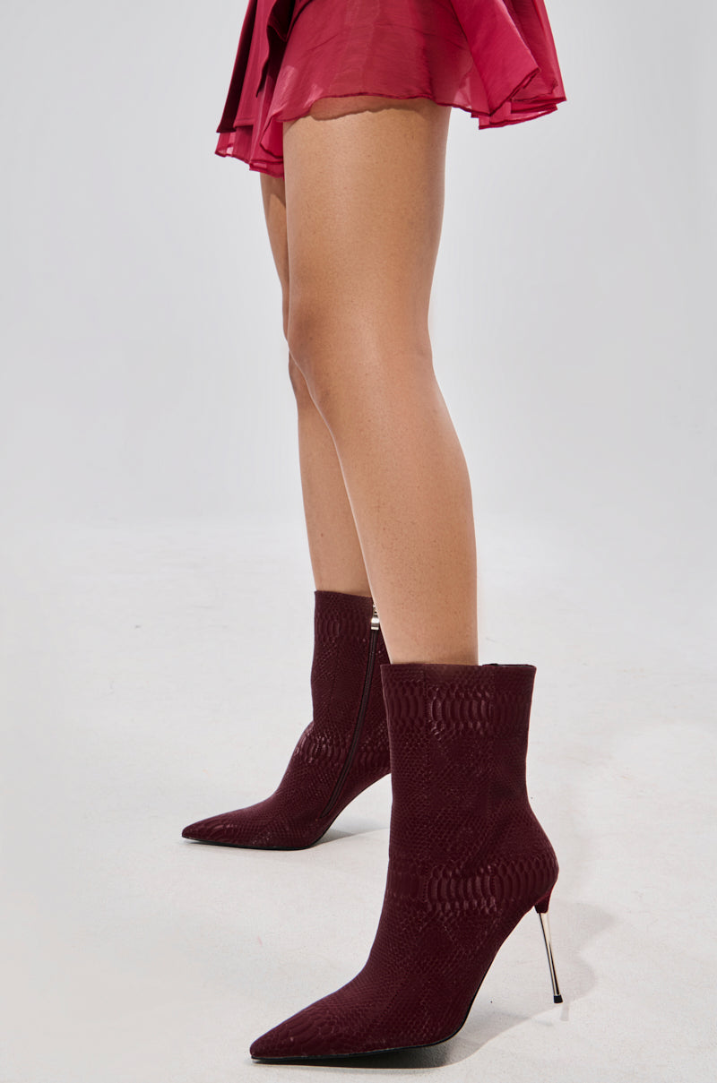 A person stands against a light grey background, wearing a short dark red skirt and the AZALEA WANG CALEAN BURGUNDY BOOTIE—faux leather ankle boots with pointed toes and stiletto heels.
