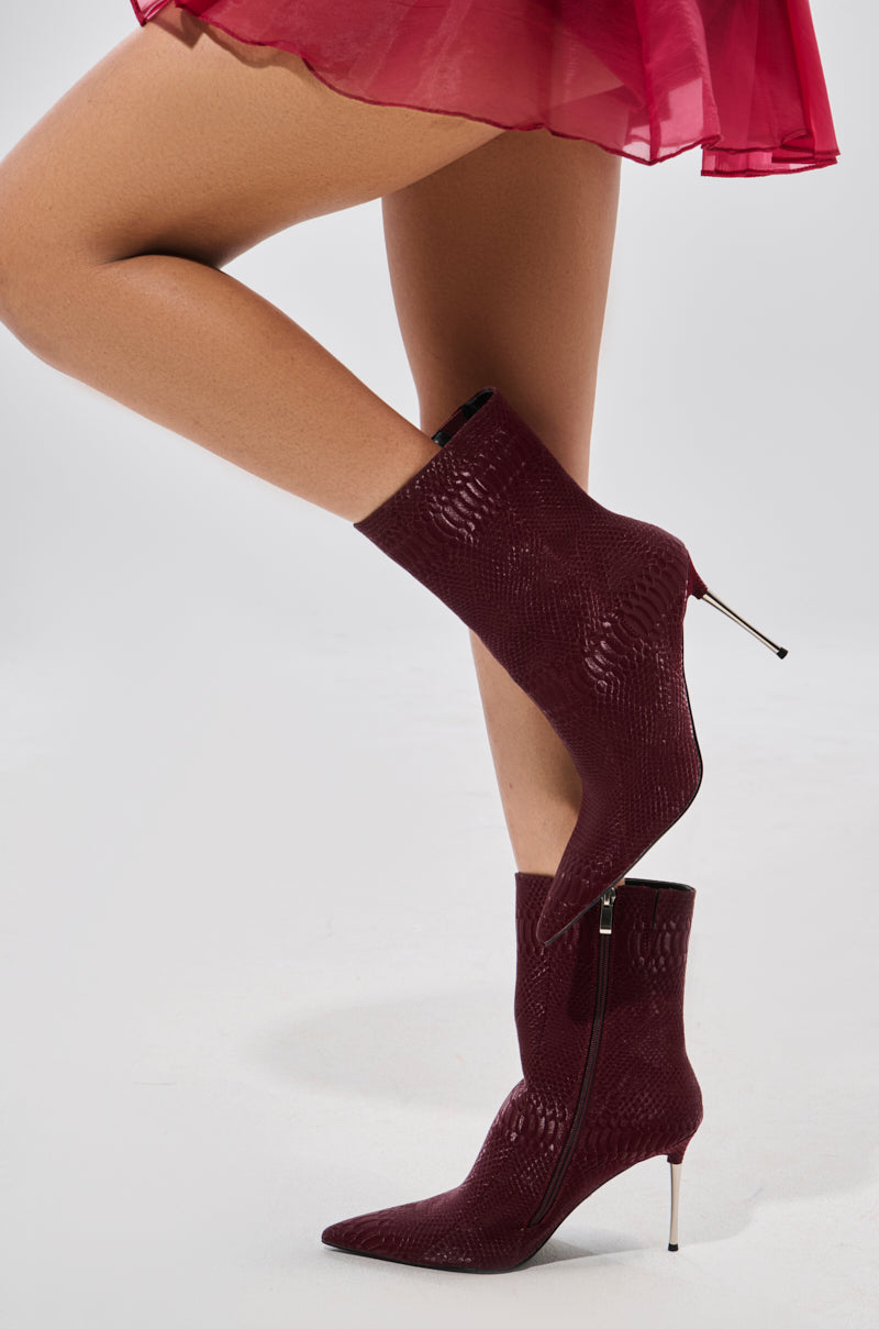 A person in a sheer red skirt and AZALEA WANG CALEAN BURGUNDY BOOTIE with a textured snakeskin pattern, pointed toes, and stiletto heels stands with one leg crossed over the other.