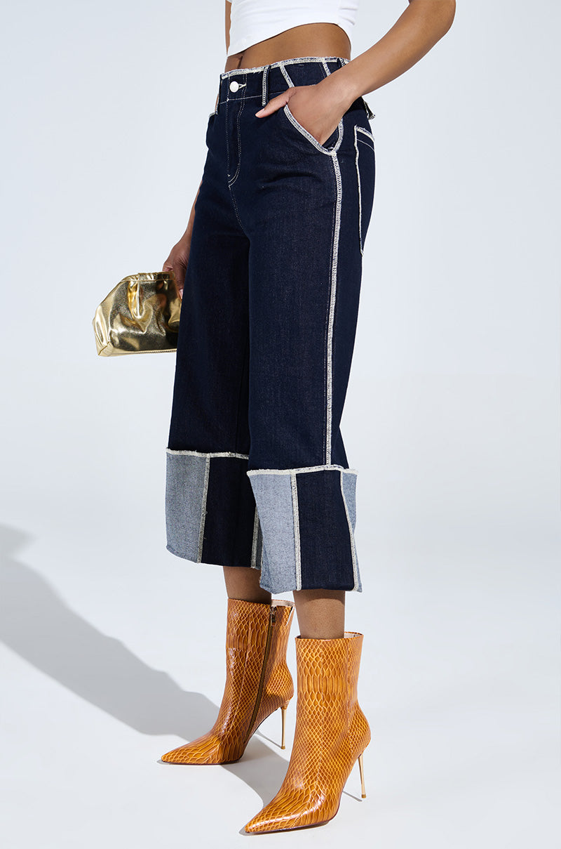 A person in dark wide-leg jeans and a white top holds a small metallic bag. Their look is completed with AZALEA WANG CALEAN ORANGE STILETTO BOOTIE in a snakeskin pattern. The background is light-colored.