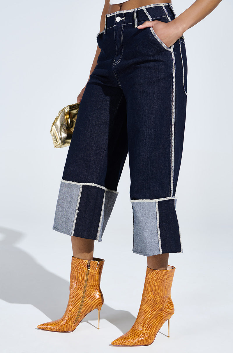 A woman showcases wide-leg denim pants with wide cuffs, paired with AZALEA WANG CALEAN ORANGE STILETTO BOOTIE and a metallic gold purse. Her style is elevated by the chic texture of faux leather in a minimalistic setting.