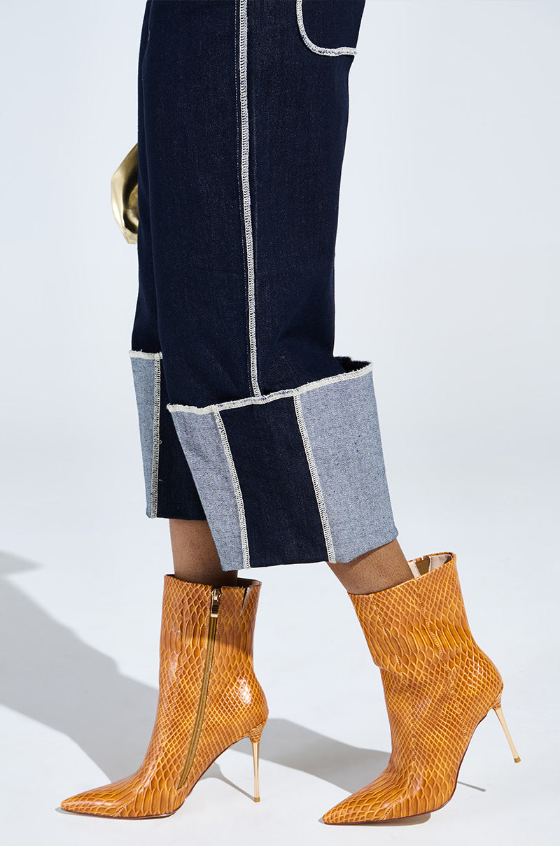 A person is wearing dark blue denim pants with light blue cuffed hems and AZALEA WANG CALEAN ORANGE STILETTO BOOTIES, featuring a snakeskin pattern with pointed toes and slim high heels. The background is plain white, with a hint of a gold glove visible.