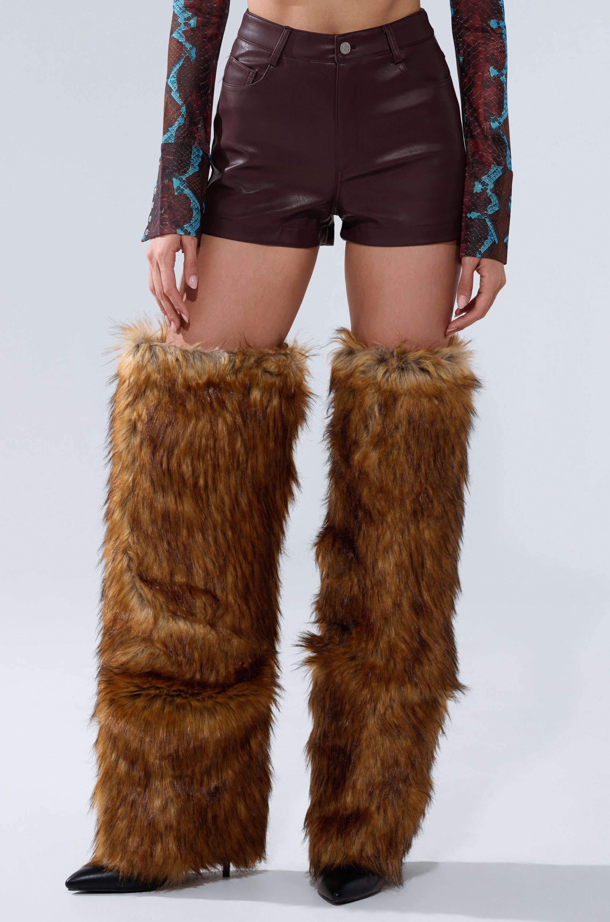 A person wears CALLIE FAUX LEATHER SHORT IN BURGUNDY with large, furry brown leg warmers over black pointed shoes, and a patterned long-sleeve top visible from the elbows down.