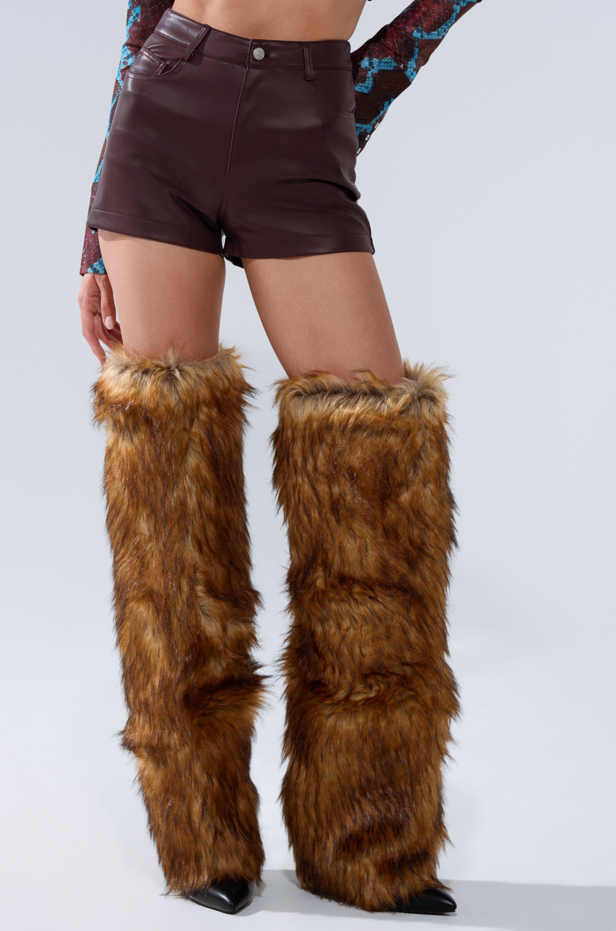 A person stands against a plain light background wearing CALLIE FAUX LEATHER SHORT IN BURGUNDY, a patterned long-sleeve top, and tall brown faux fur leg warmers over black shoes.