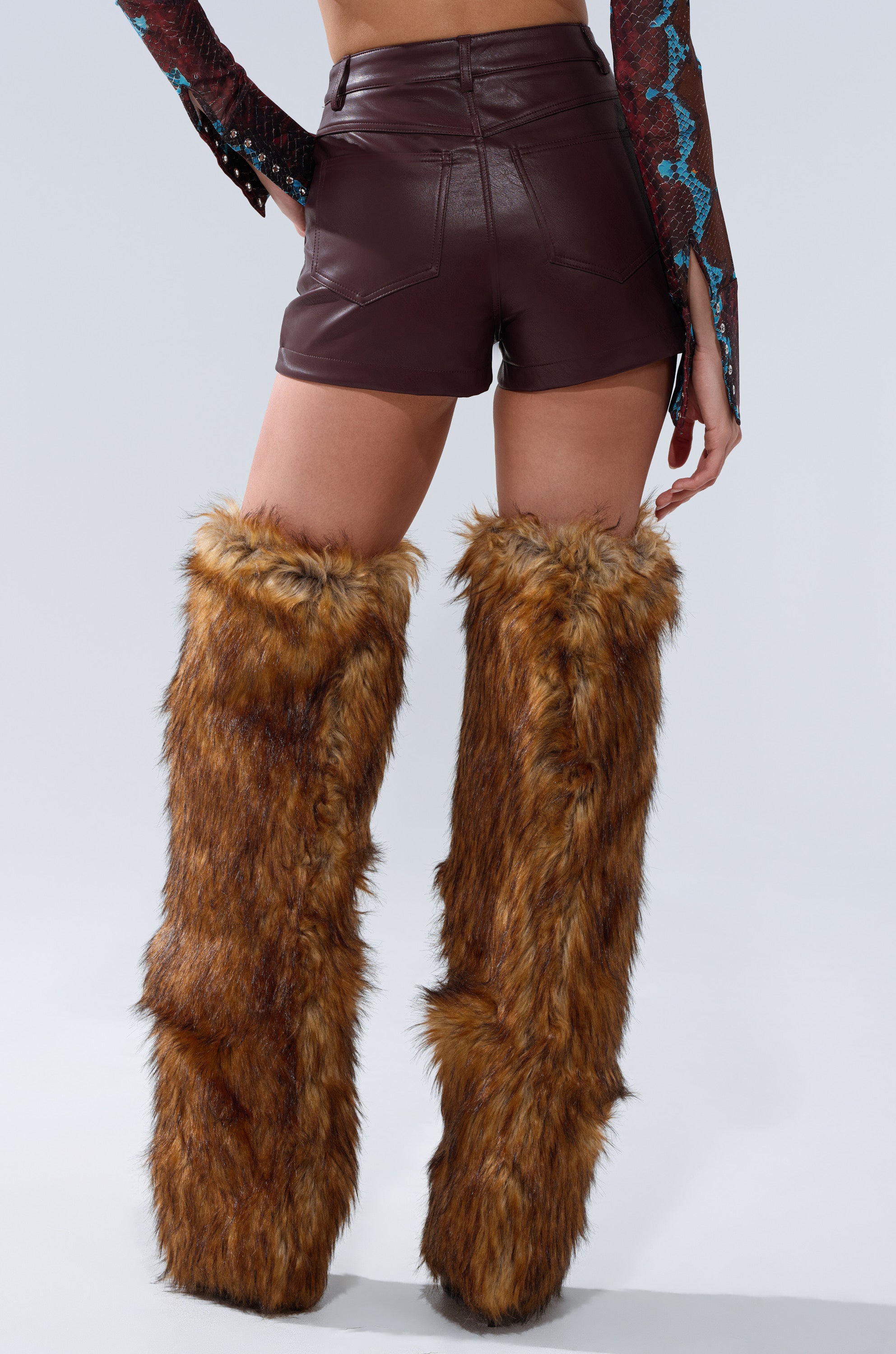 A person wearing CALLIE FAUX LEATHER SHORT IN BURGUNDY paired with tall furry brown leg warmers and platform shoes, shown from the back against a plain light background.
