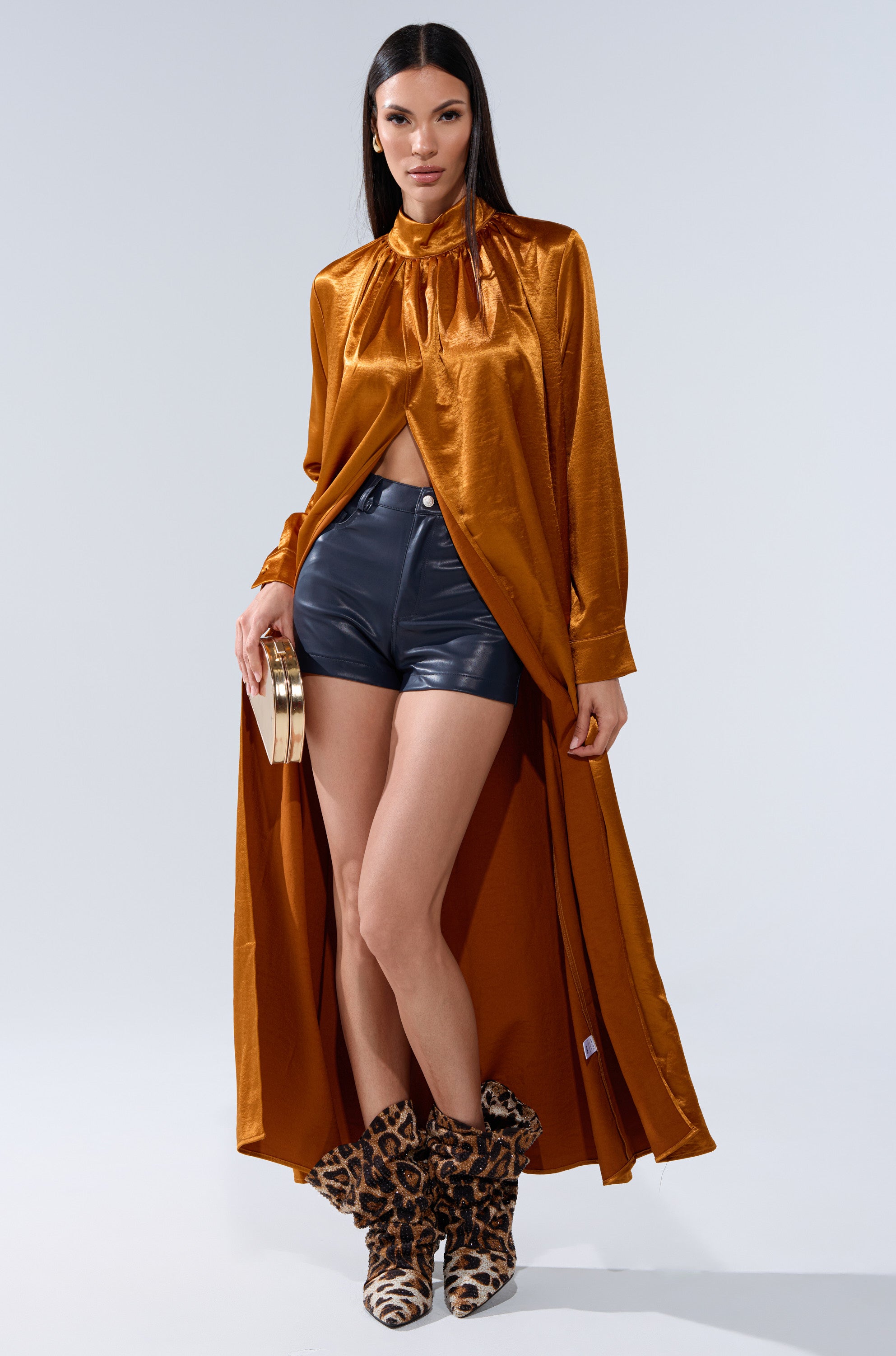 A woman poses confidently in a shiny orange long-sleeve coat, CALLIE FAUX LEATHER SHORT IN NAVY, leopard print boots, and a small gold clutch against a plain light background.