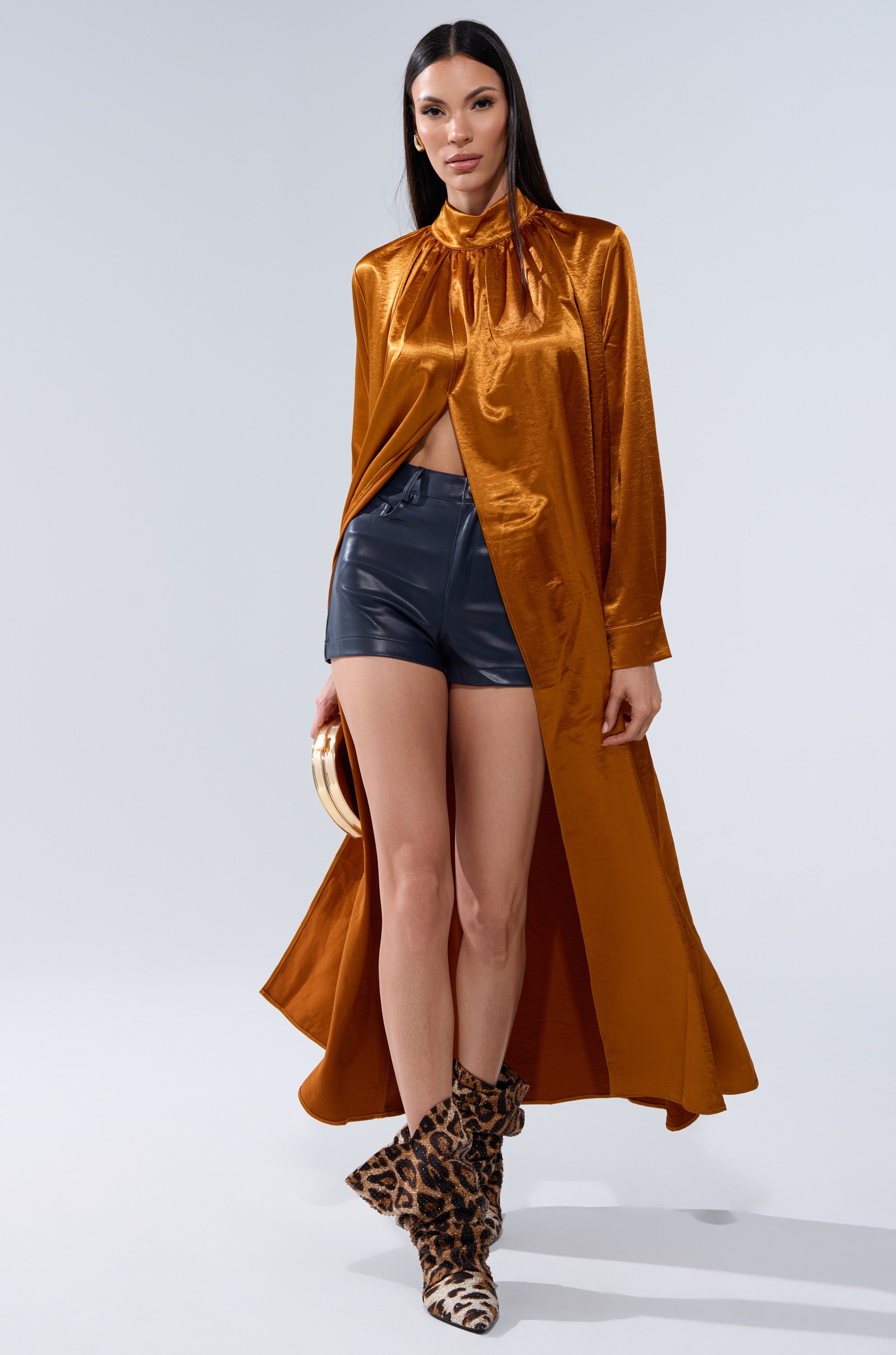 A woman wearing a shiny orange long-sleeve high-neck coat, CALLIE FAUX LEATHER SHORT IN NAVY, and leopard print boots stands against a light background, holding a small round bag.