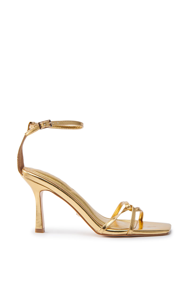 The AKIRA CALSITE GOLD STILETTO SANDAL is a gold faux leather shoe featuring thin straps, an ankle buckle, and an open square toe. Its sleek, elegant design with a moderately high heel stands out against a plain white background.