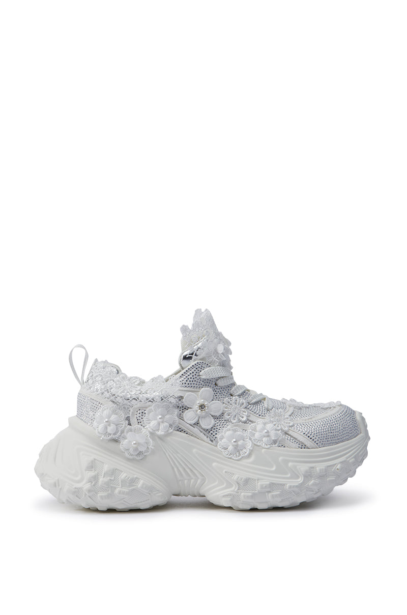 The AZALEA WANG CANISH WHITE EMBELLISHED SNEAKER features a unique and artistic flair with intricate floral lace detailing on white platform sneakers. The chunky textured sole is complemented by a mesh fabric beneath the lace, enhanced with rhinestone embellishments for an eye-catching appearance that captures attention.
