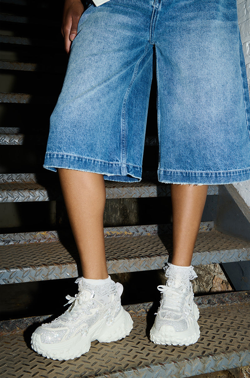 A person wearing baggy denim shorts and the AZALEA WANG CANISH White Embellished Sneakers with frilly socks is standing on a metal staircase.
