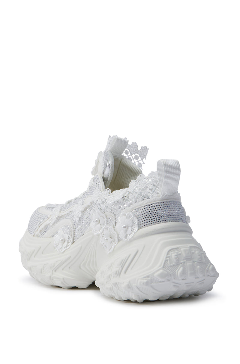 The AZALEA WANG CANISH WHITE EMBELLISHED SNEAKER is adorned with lace and rhinestones on the sides, showcasing thick, textured platform soles and delicate floral detailing from the back angle.