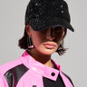 A person wearing the "PUT IT IN MOTION RHINESTONE BASEBALL CAP" in black, embellished with sequins and rhinestones, paired with large silver hoop earrings and a pink jacket featuring black accents, stands against a plain gray background. They have a serious expression and a septum piercing.