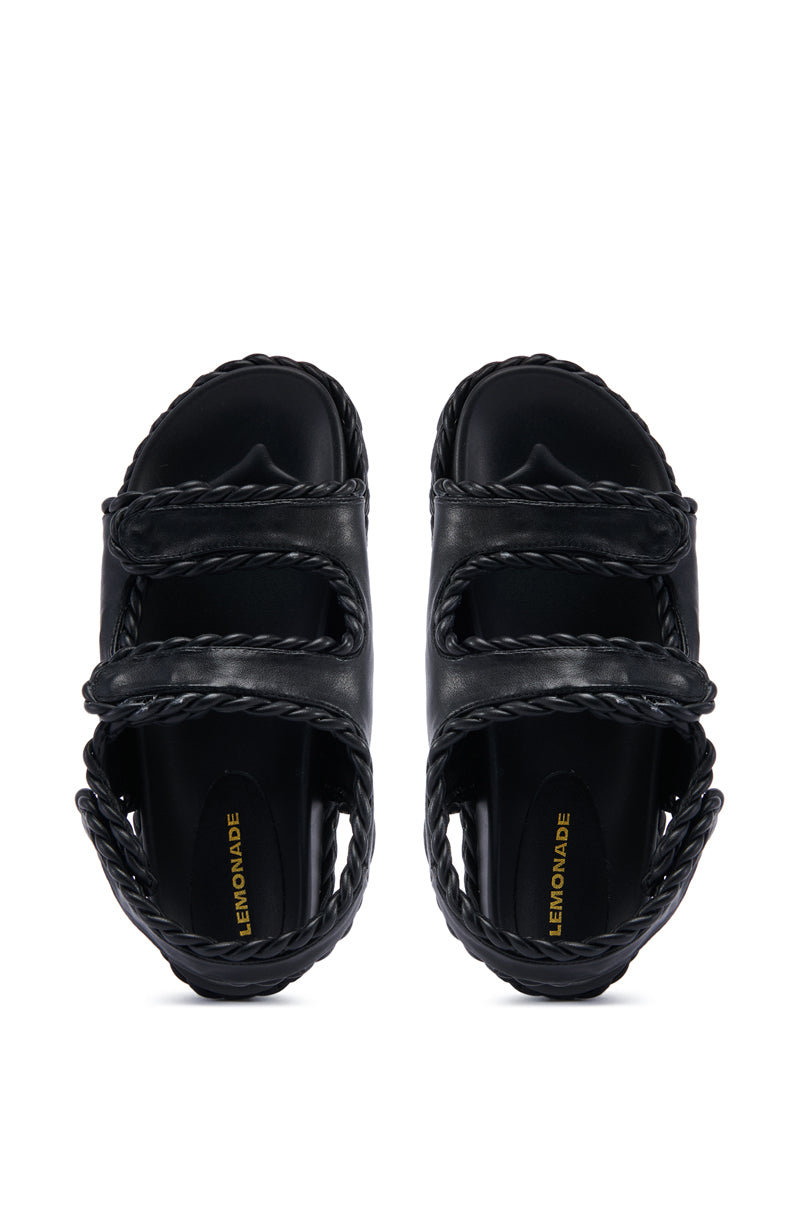 The CAPE COD BLACK BRAIDED SANDAL with a faux leather upper and velcro straps is displayed. This minimalist summer shoe features "LEMONADE" printed in yellow on the footbed. Viewed from the top, both sandals are shown side by side, highlighting their intricate braided details.