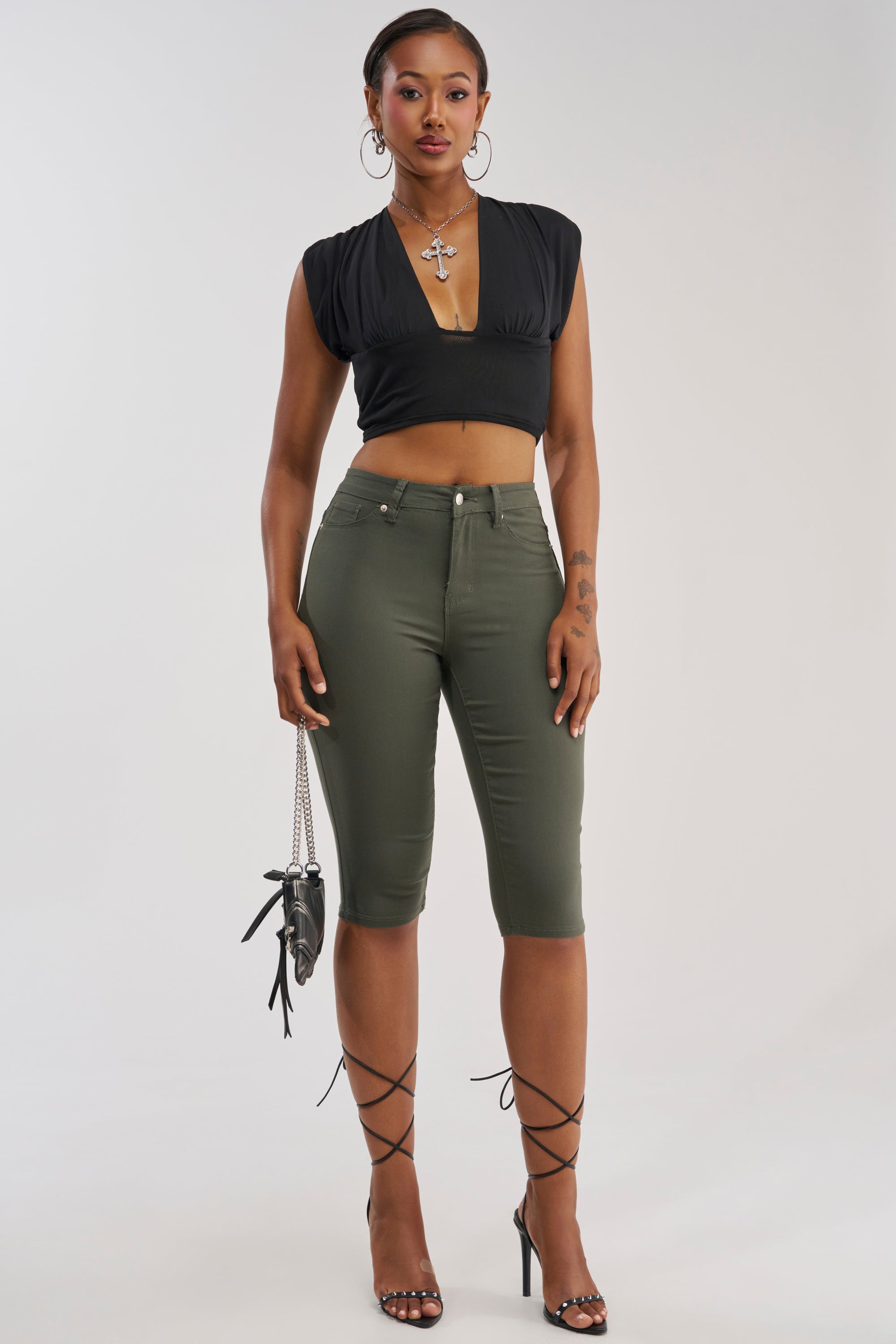 A woman stands confidently against a plain background wearing the DENIM MUSE CAPRI in olive green, paired with a black crop top and lace-up heels. She accessorizes with a small black purse, hoop earrings, and a cross necklace, with her hair styled back.