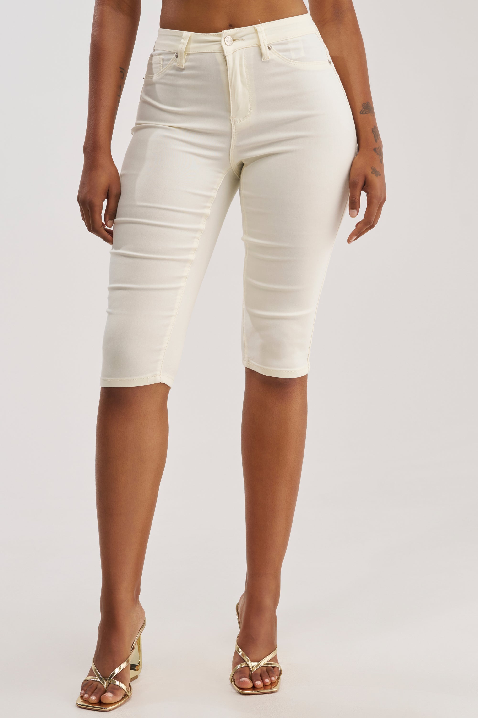 A person is shown wearing the Denim Muse Capri in White—high-waisted, knee-length capri shorts—paired with gold open-toe heels, standing against a plain white background. Only the lower half of the body is visible.