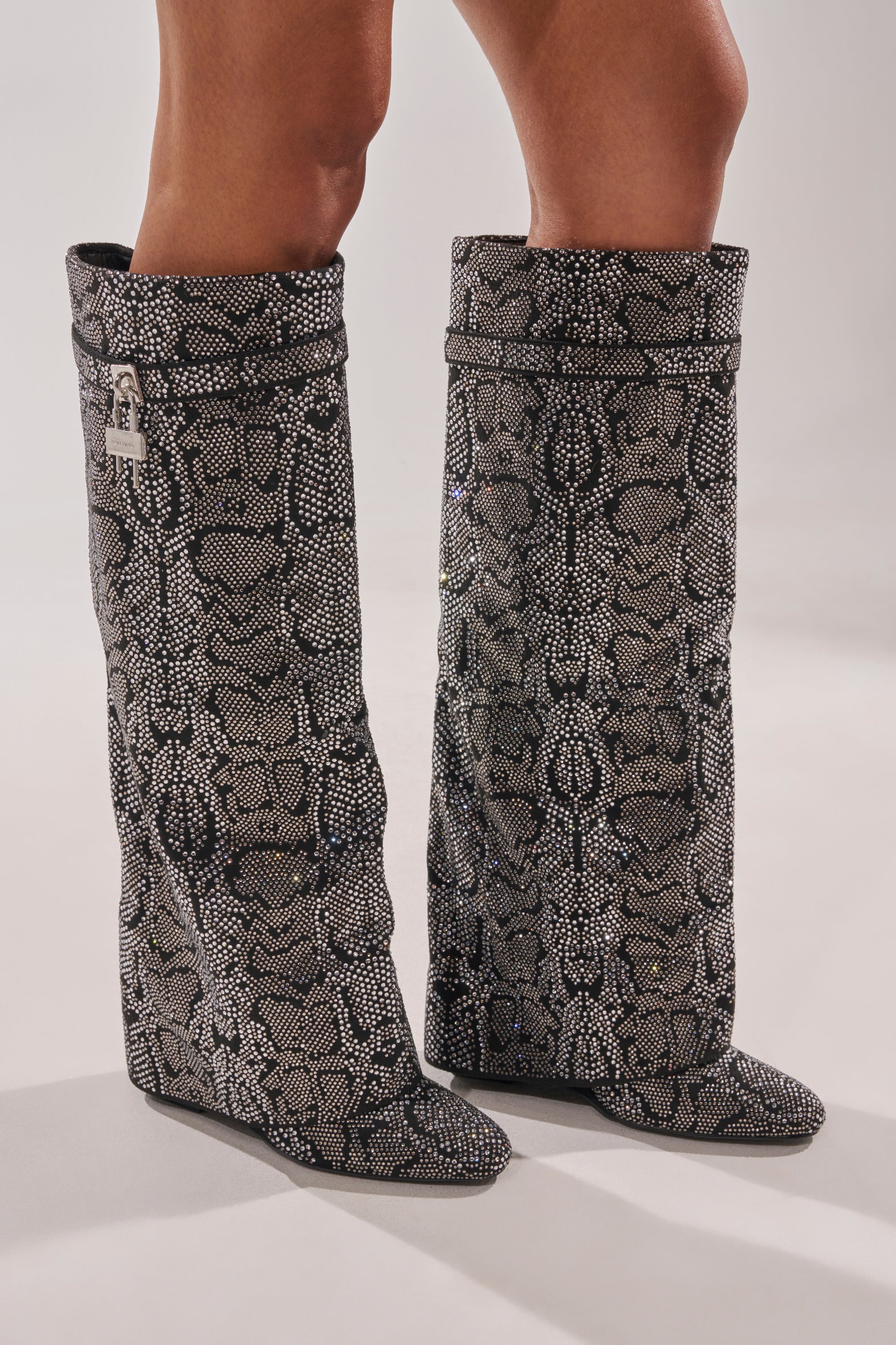 A person wears the AZALEA WANG CAPRICORN RHINESTONE SNAKE PATTERN WEDGE FOLD OVER BOOT—knee-high, black and gray snakeskin with a loose, slouchy fit, pointed toes, and wedge heels—while standing on a light surface. 0