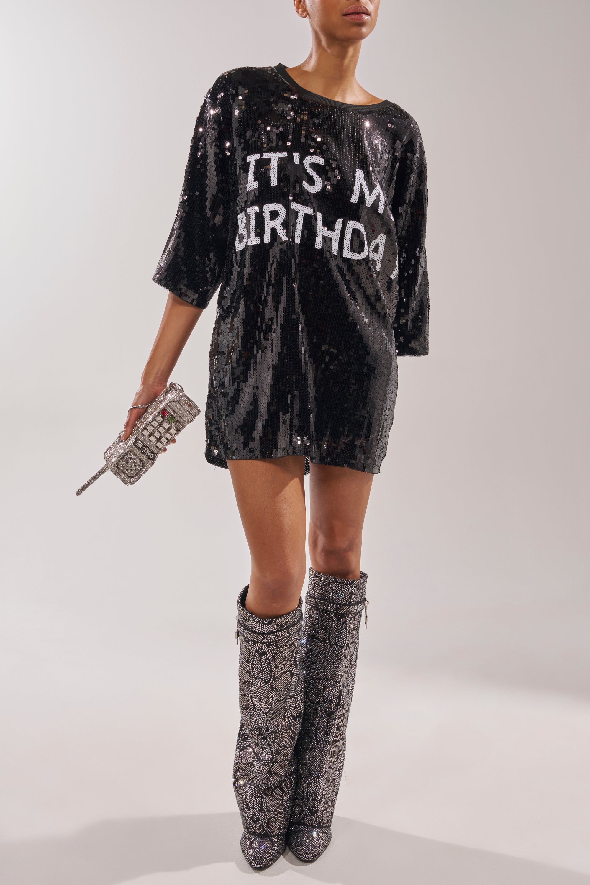 A person in a black sequin "IT’S MY BIRTHDAY" dress holds a rhinestone phone-shaped clutch and wears AZALEA WANG CAPRICORN RHINESTONE SNAKE PATTERN WEDGE FOLD OVER BOOTS. Face not shown. 1