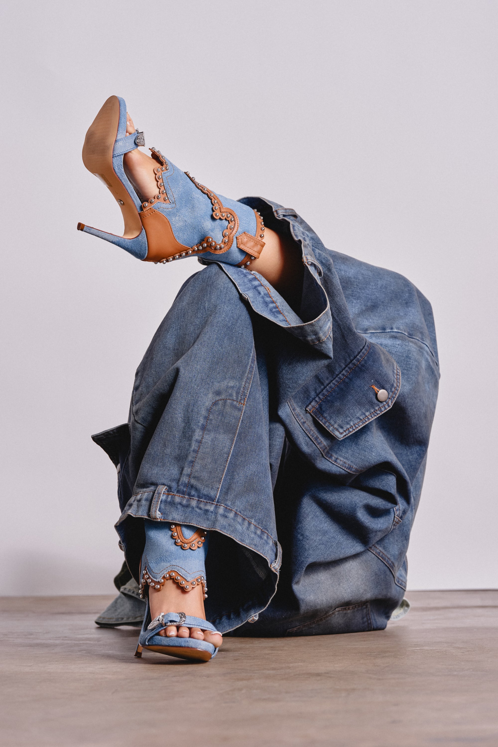 Wearing oversized denim, a person sits with one leg raised, showcasing the AZALEA WANG CAPRIZIA DENIM WESTERN NOVELTY STUDDED STILETTO SANDAL—blue and tan open-toe heels with gold embellishments, set against a plain background. 1