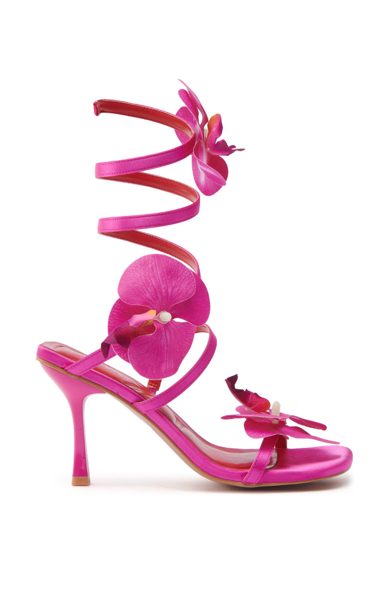 The AZALEA WANG CARRINGTON FUCHSIA FLORAL COIL SANDAL features a satin upper with a bright pink finish, large orchid flower accents, wraparound ankle straps, and a slender mid-height stiletto heel.