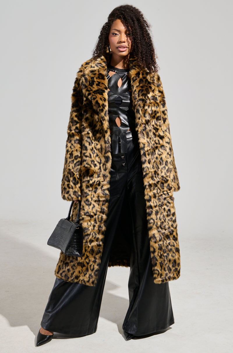 A woman wearing the CATTY FAUX FUR LEOPARD COAT draped over a black, cut-out leather top and wide-leg pants accessorizes with a small black handbag while posing against a plain light gray background.