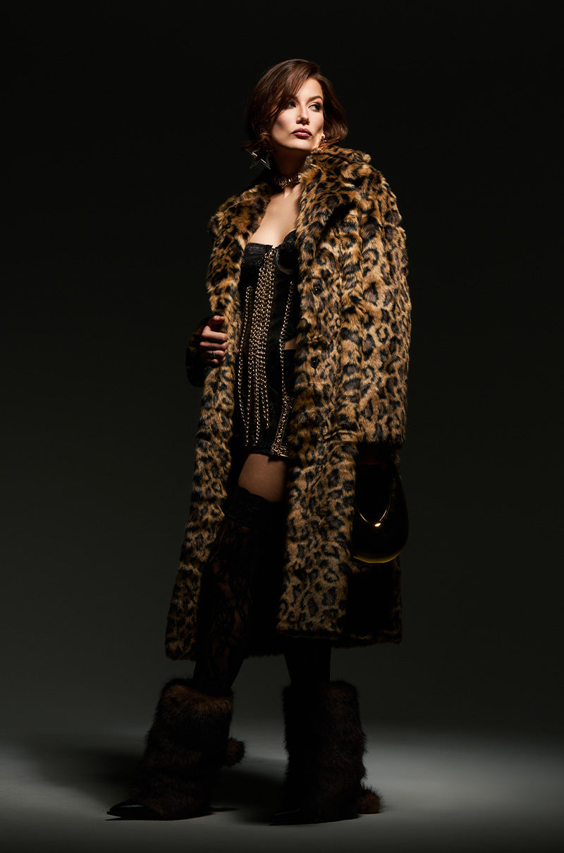 A woman poses against a dark background, draped in the CATTY FAUX FUR LEOPARD COAT over a black outfit with gold chain accents. She carries a small purse and sports flatform boots embellished with faux fur, radiating a glamorous and confident vibe.