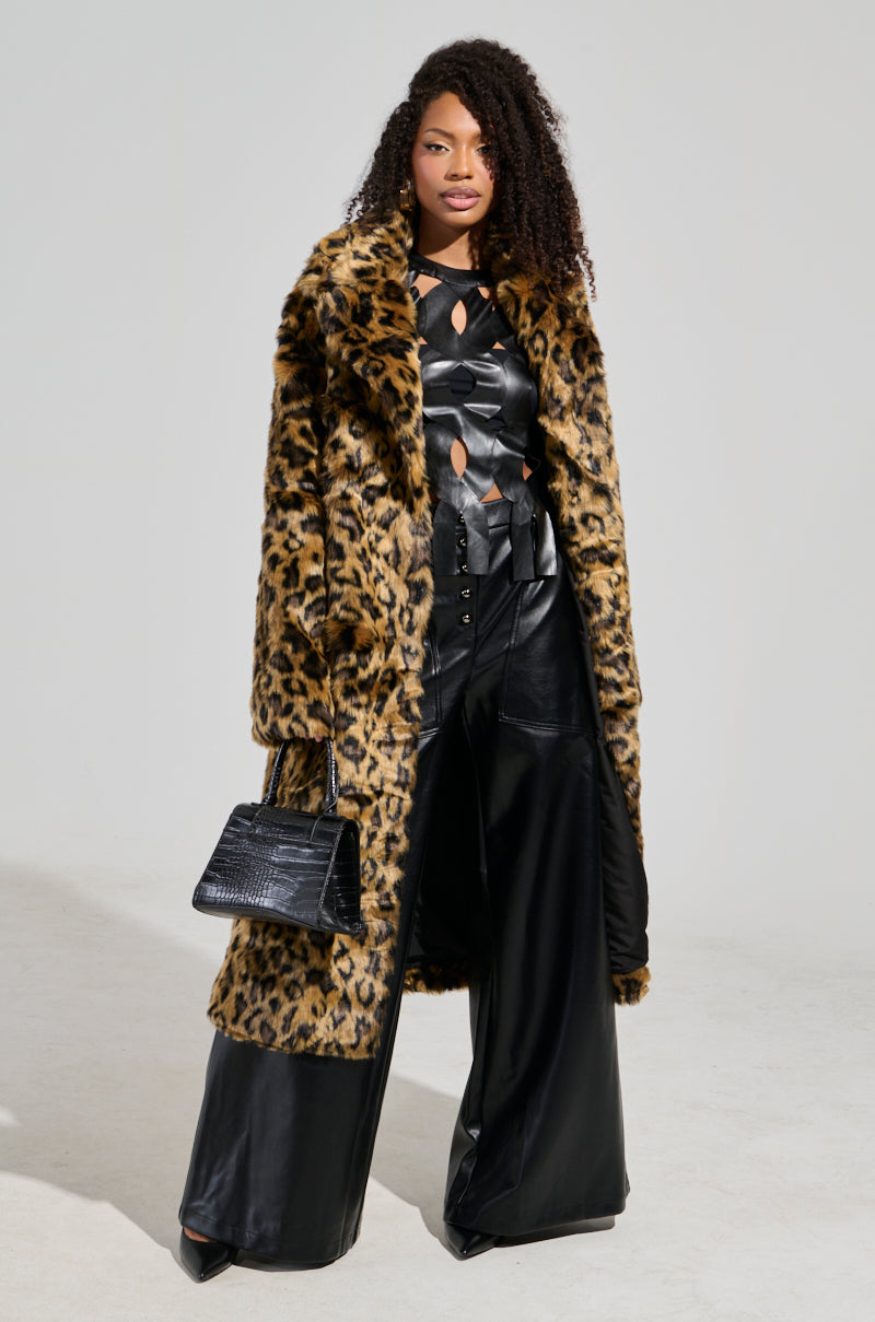 A woman stands confidently, her curly hair cascading over the CATTY FAUX FUR LEOPARD COAT draped over a sleek black leather outfit. She accessorizes with a black handbag and eye-catching flatform boots, striking a bold pose against the plain backdrop.