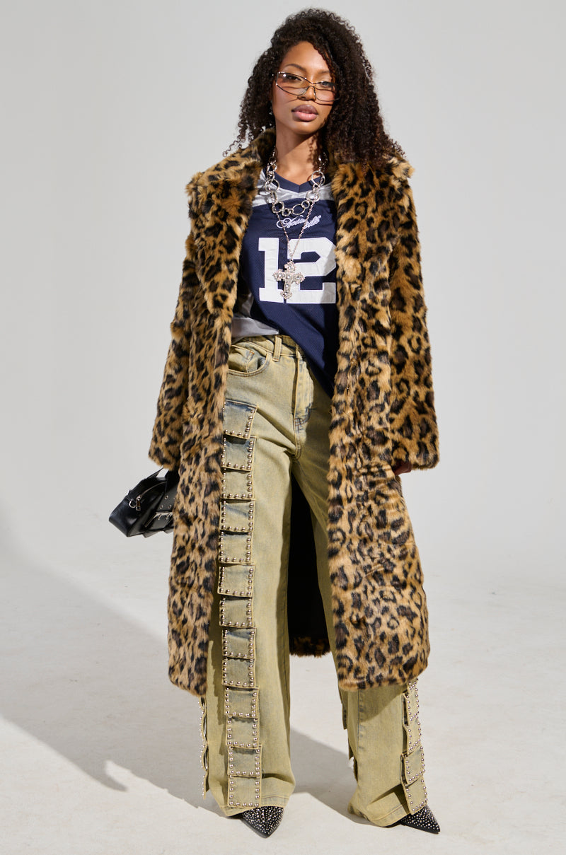 A person exudes confidence in the CATTY FAUX FUR LEOPARD COAT layered over a navy shirt with "12" on it, complemented by wide-leg khaki pants adorned with strap details. They accessorize with a small black bag and wear curly hair, sunglasses, and flatform boots.