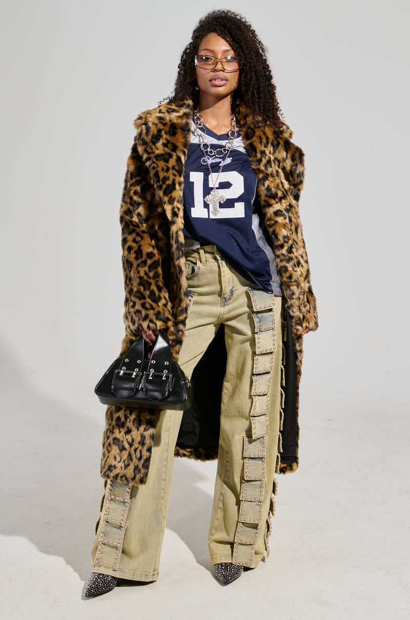 Wearing the CATTY FAUX FUR LEOPARD COAT, an individual poses with a blue jersey featuring the number 12. They pair this with beige pants featuring side detailing and carry a black handbag. Their curly hair is styled, and they accessorize with glasses, a necklace, and pointed shoes.