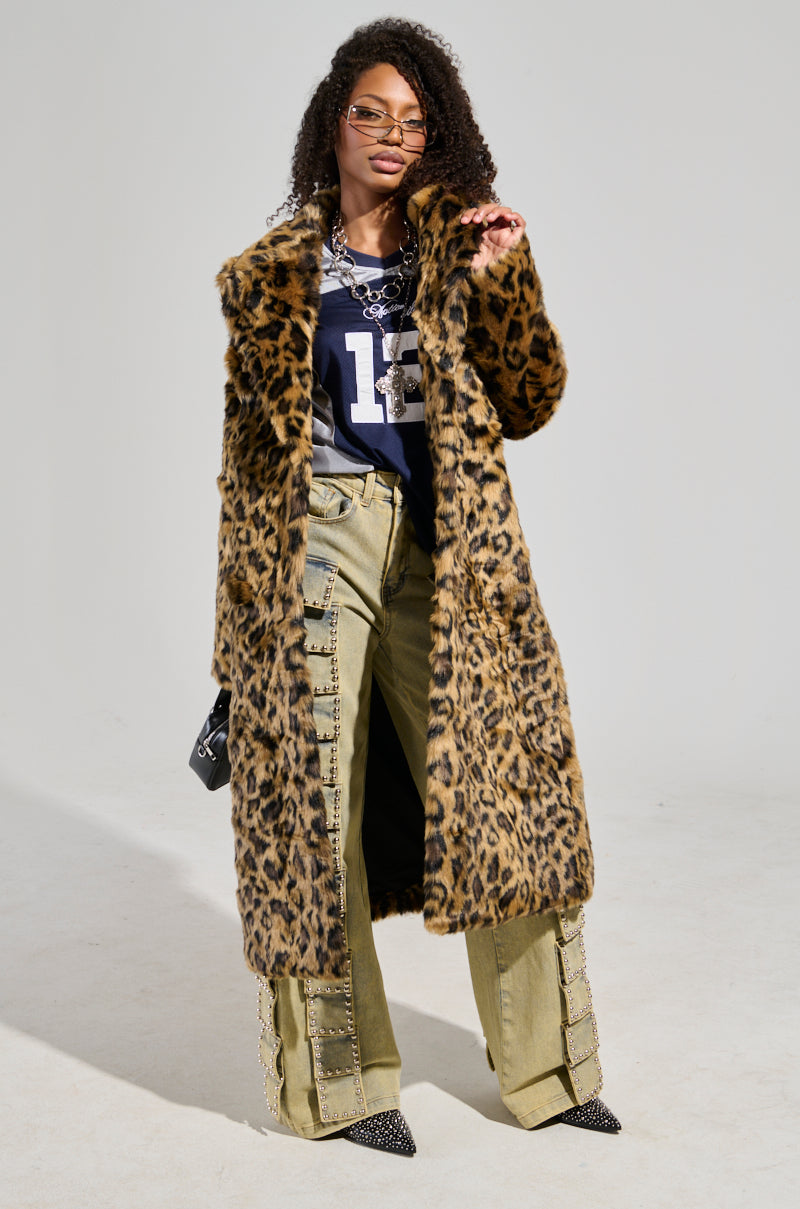 A person stands confidently in a striking CATTY FAUX FUR LEOPARD COAT over a navy shirt and green pants with embellishments. They accessorize with glasses, a necklace, and flatform boots while holding a black bag against a plain white background.