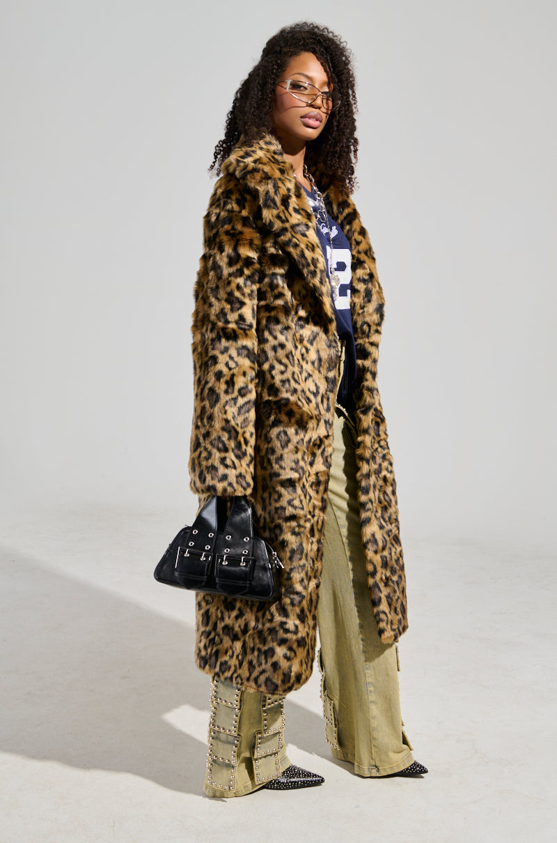 A person with curly hair stands confidently in the CATTY FAUX FUR LEOPARD COAT, paired with a blue shirt and tan pants featuring chain details. Completing the look with flatform boots and sunglasses, they carry a black handbag against a plain background.