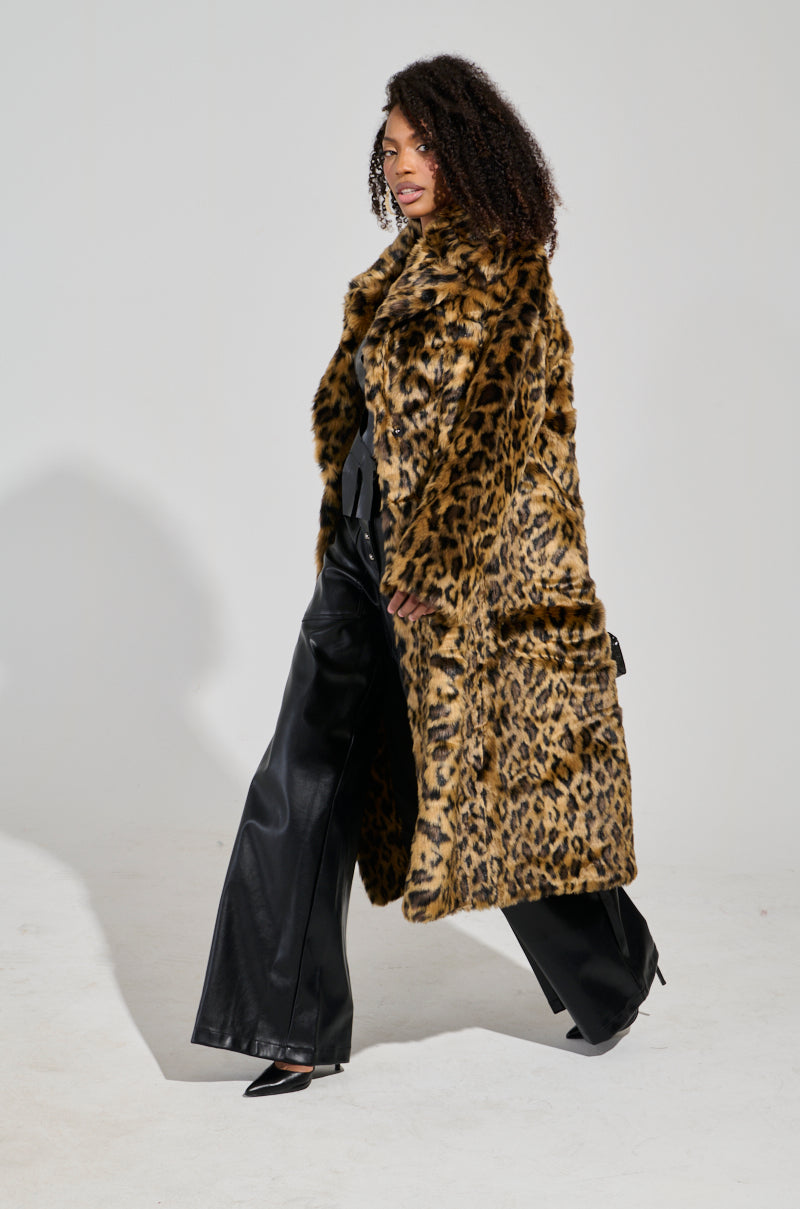 Striding confidently indoors against a plain background, a person with curly hair showcases the CATTY FAUX FUR LEOPARD COAT, complemented perfectly by black wide-leg trousers and chic flatform boots.