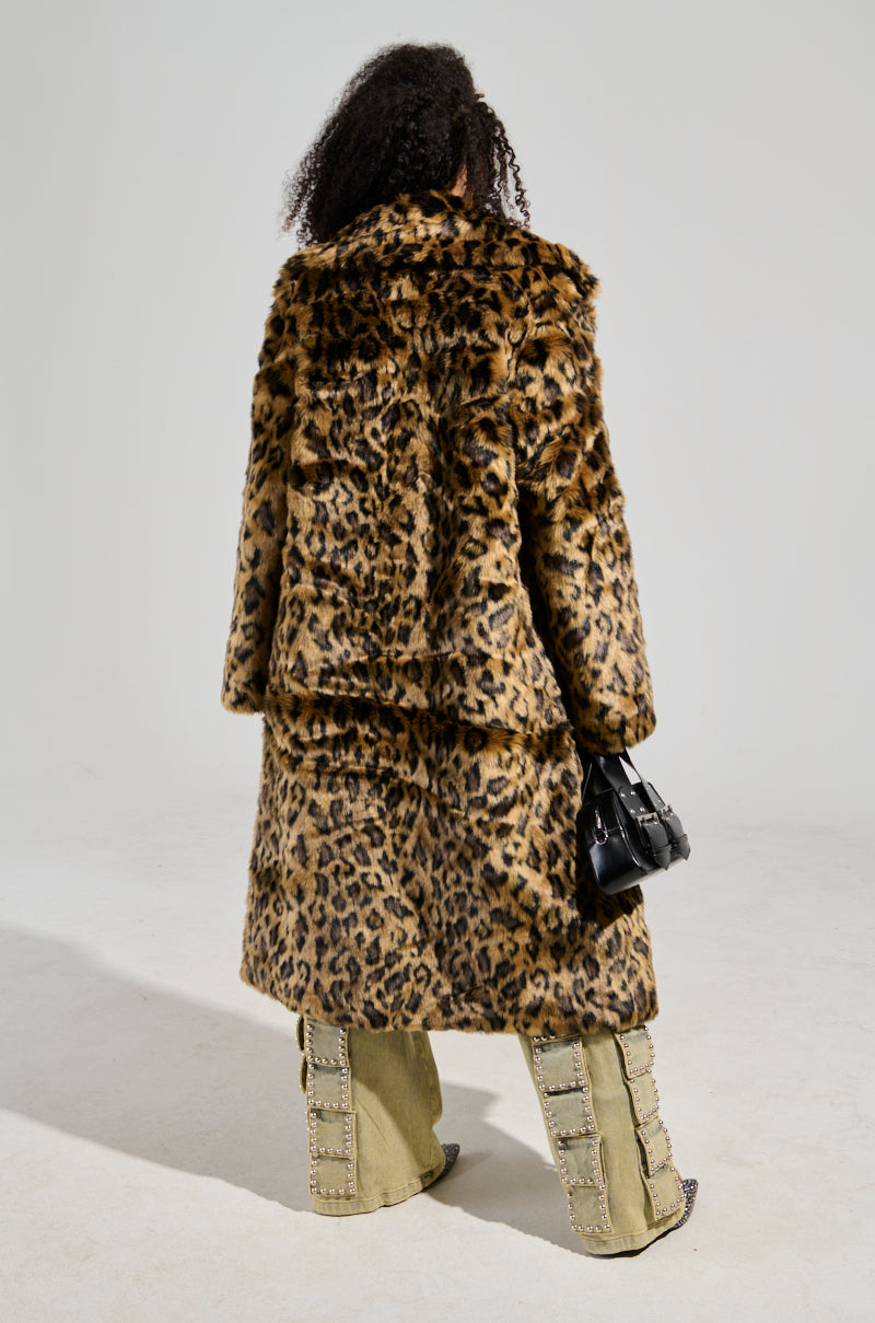 A person with curly hair is wearing the CATTY Faux Fur Leopard Coat paired with beige flared pants adorned with embellishments. They complete the ensemble with chic flatform boots and hold a small black handbag while facing away from the camera.