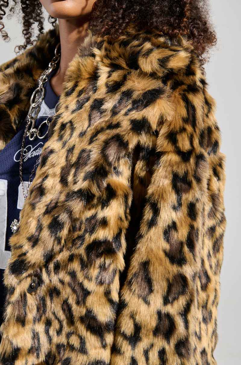 A close-up of a person wearing the CATTY FAUX FUR LEOPARD COAT, paired with flatform boots. This coat features a striking, spotted pattern in shades of brown and black. The person's curly hair peeks out, complemented by a layered necklace and jersey underneath.