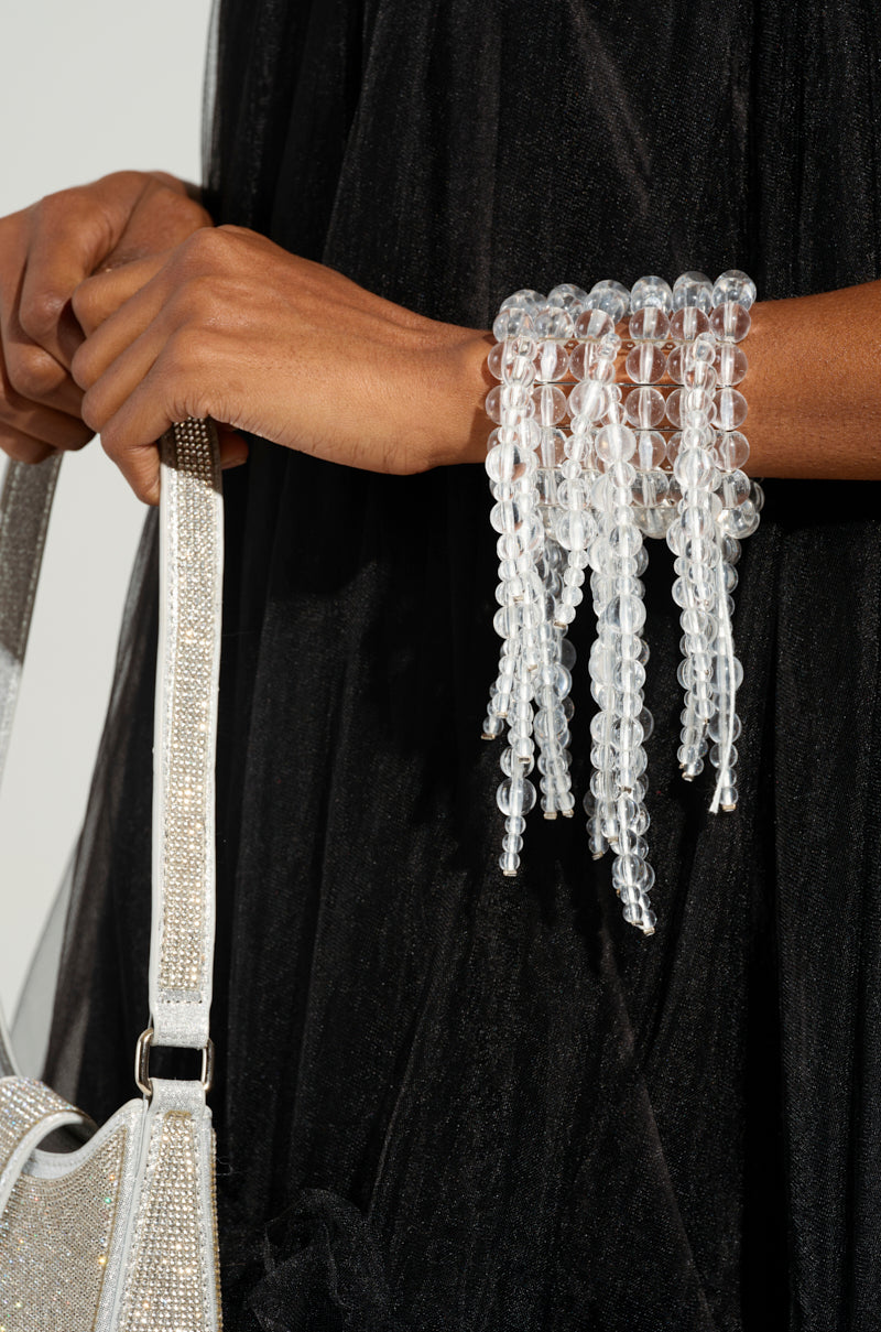 A person in a black dress holds a silver handbag, wearing the CLEAR VISION STATEMENT BRACELET, adorned with an acrylic beaded fringe of clear, dangling beads that mirror strands of bubbles, cascading elegantly down the wrist.