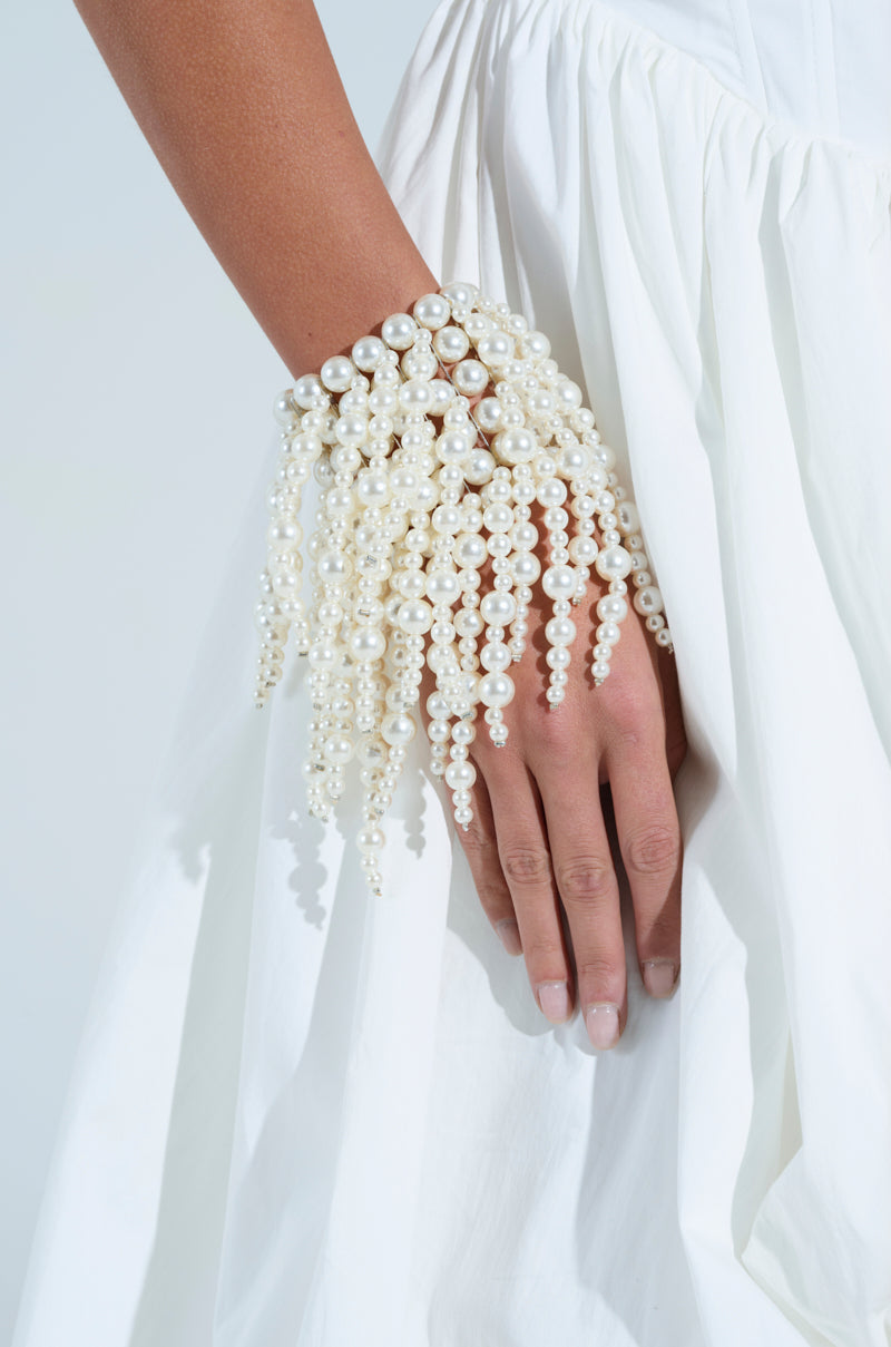 A close-up of a hand against a white dress showcases the PEARL VISION STATEMENT BRACELET, its cascading strands elegantly draping down the wrist like fringe. The bracelet exudes luxury and sophistication against a soft, neutral backdrop. 1